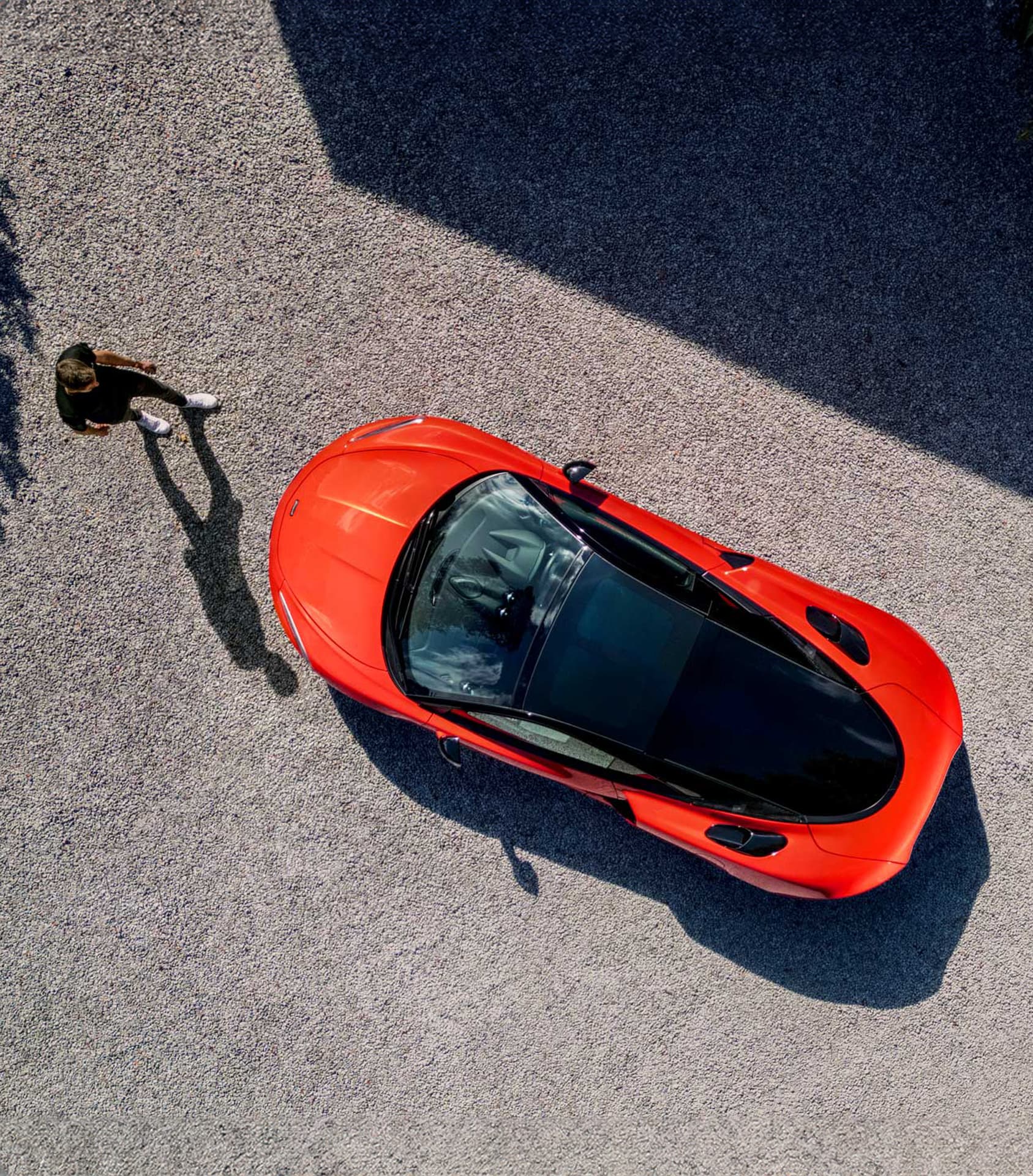 An aerial view shows a bright red McLaren GTS parked on a gravel driveway next to a person, with stark shadows.