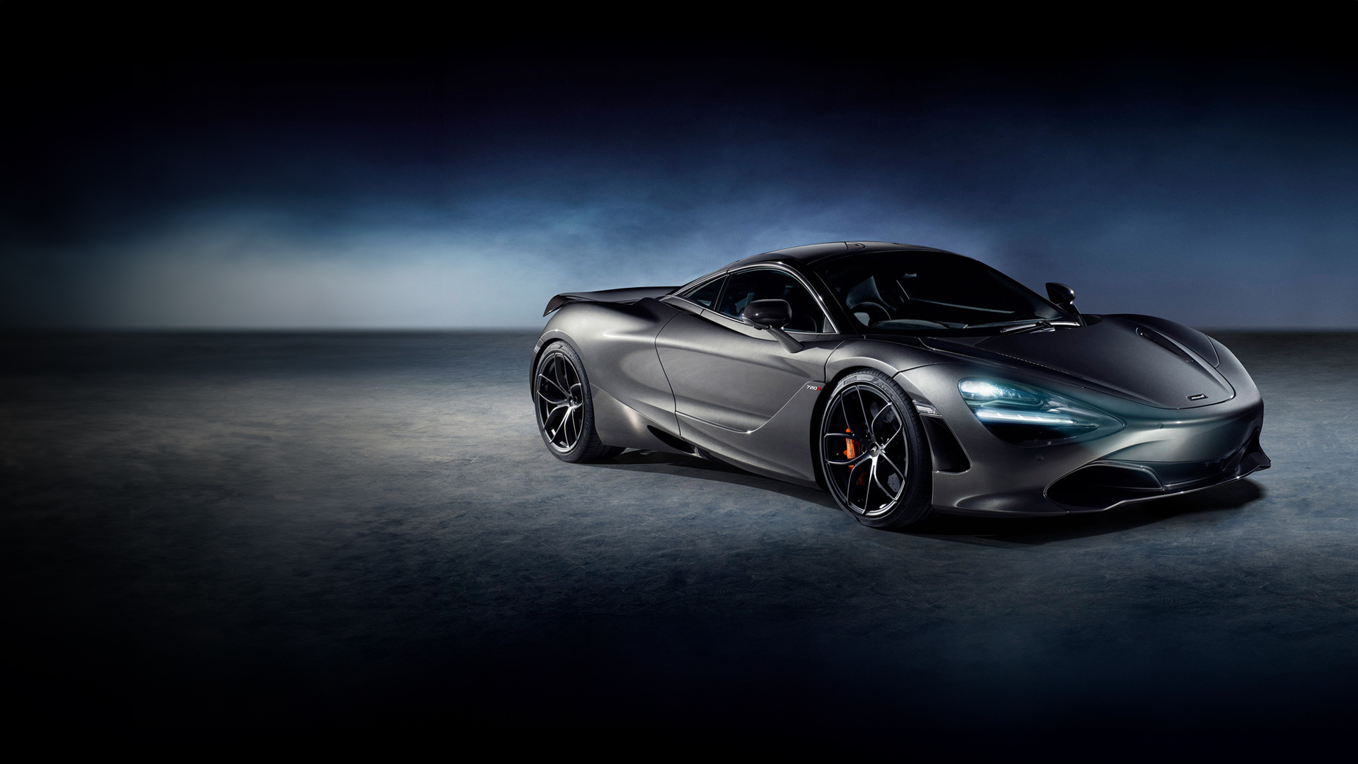 A grey McLaren 720S sports car is parked in a moody studio setting, with its sleek body lines and headlights on, highlighting the front wheel and orange brake caliper.