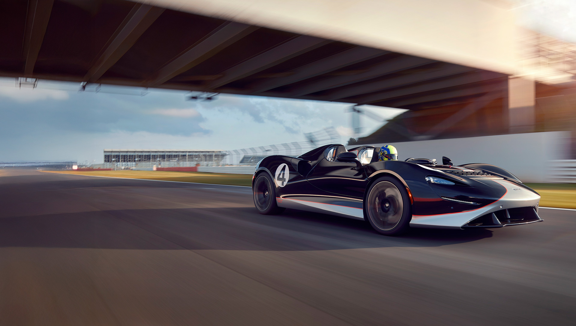 A black and white McLaren Elva roadster with the number 4 is driven on a racetrack, passing under a bridge in a dynamic action shot.