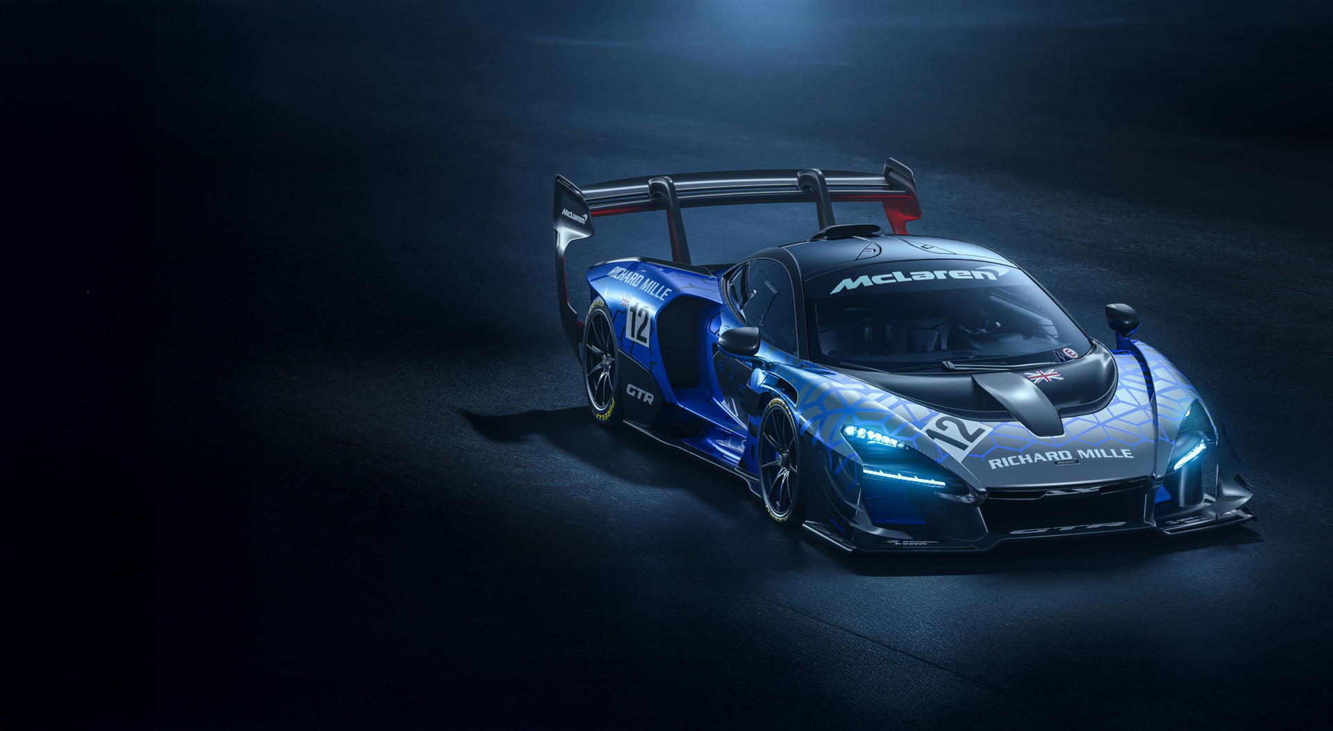 Blue patterned McLaren Senna GTR parked in a dark concrete setting with headlights on.