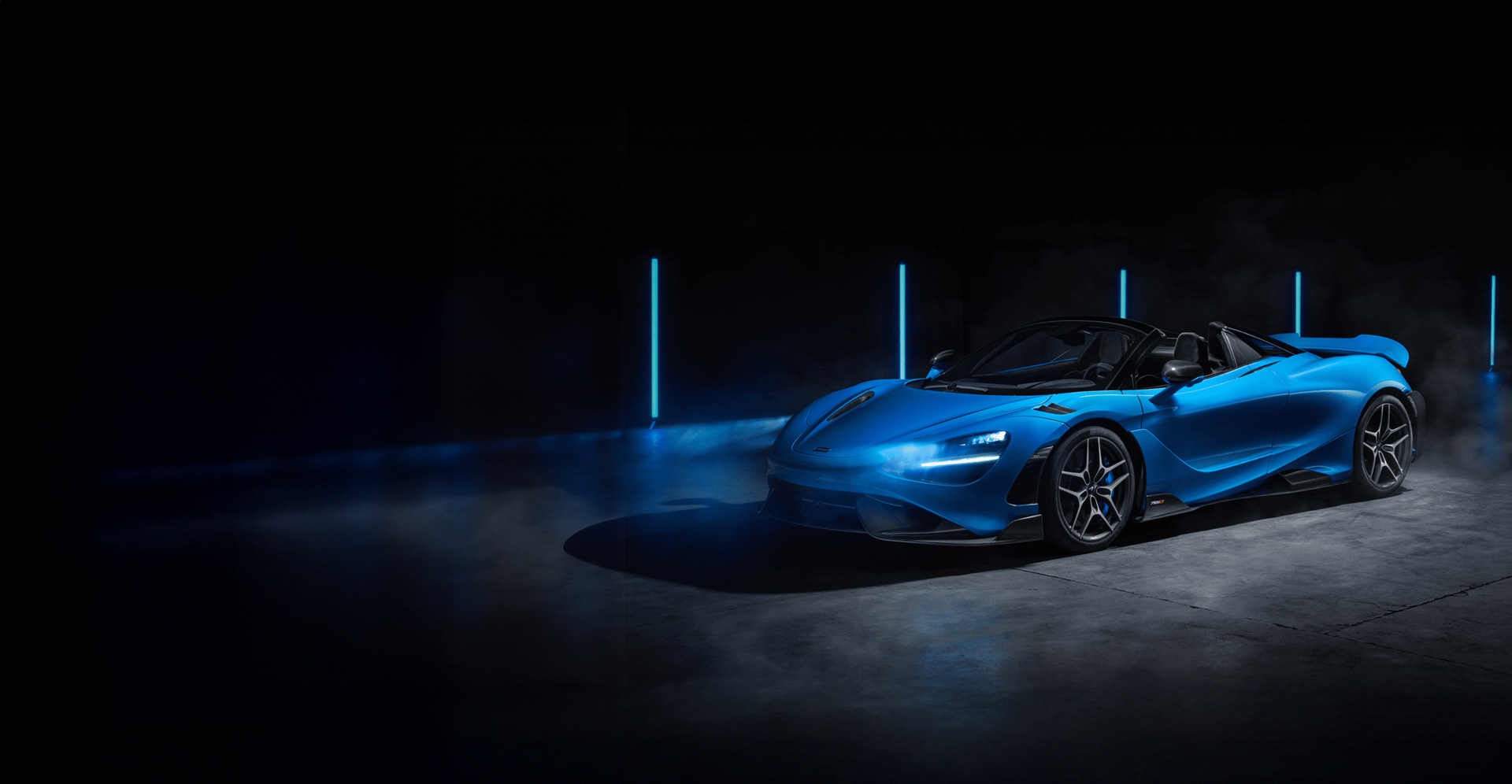 A studio shot of a McLaren 765LT Spider convertible in a bright metallic blue. The car is parked on a dark, concrete floor with its roof down.