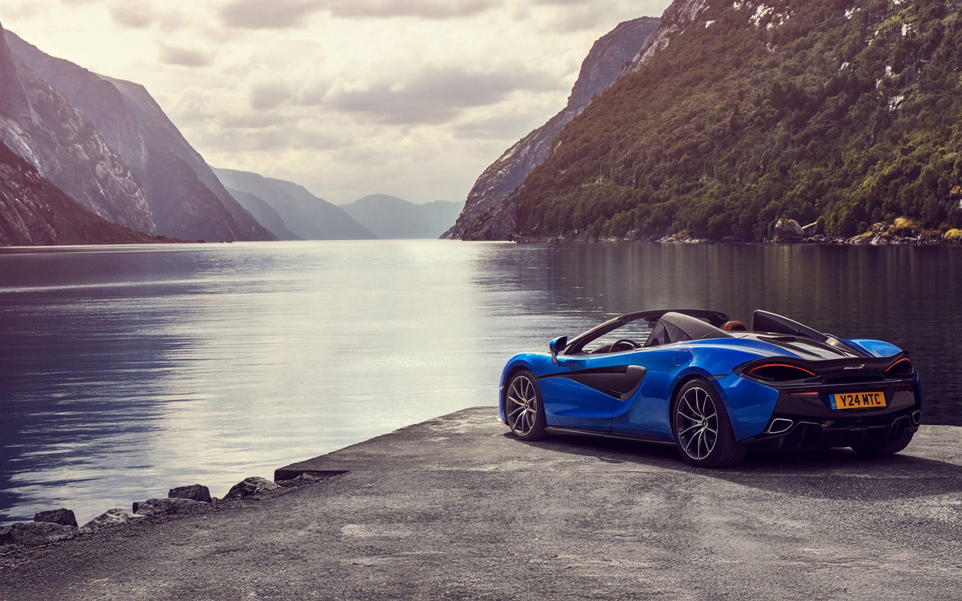 A blue McLaren 570S Spider parked on a concrete dock, overlooking a lake, surrounded by mountains covered in greenery.