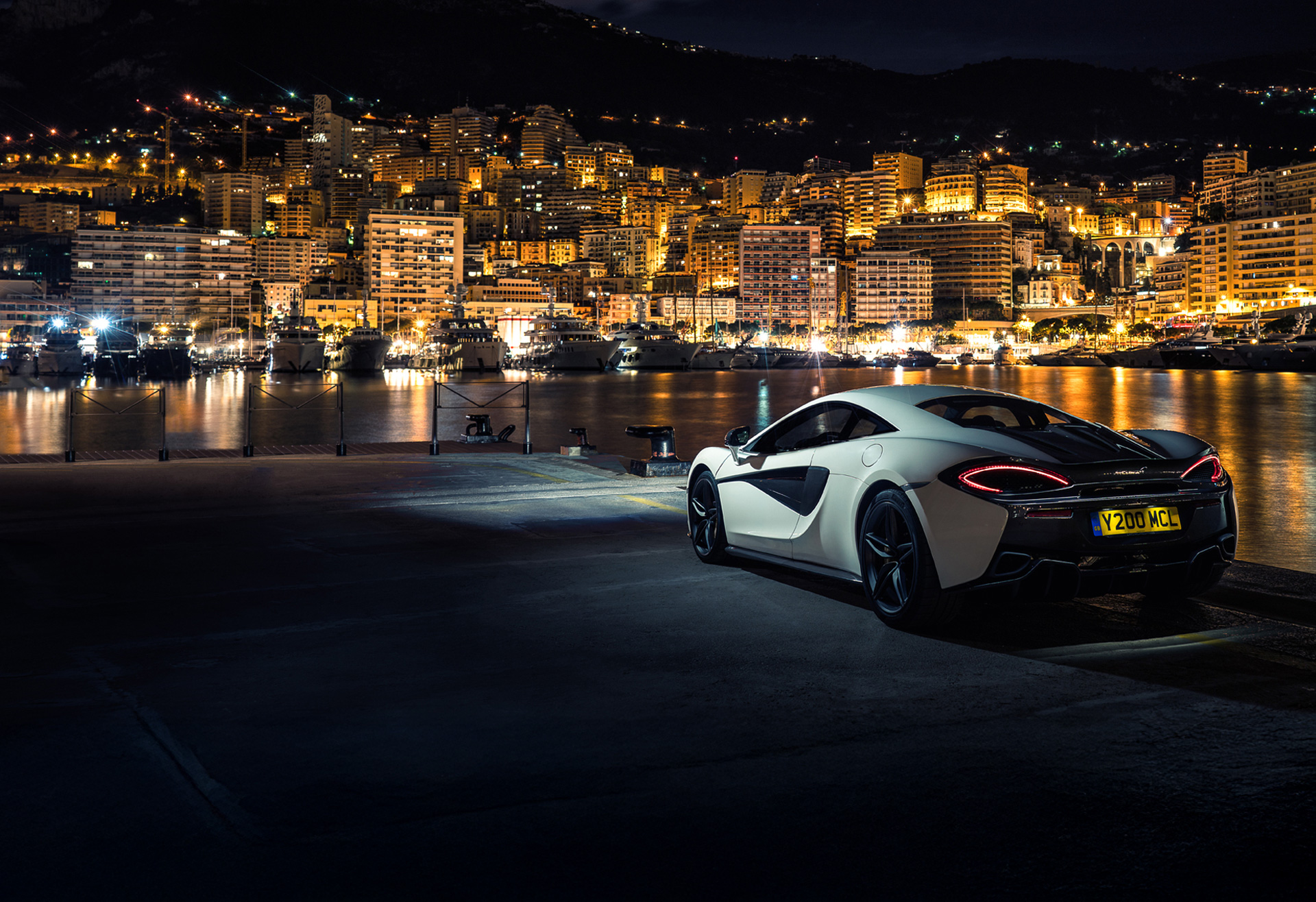 White McLaren 540C parked on the waters edge, with headlights on, overlooking Monte Carlo, Monaco lit up at night. 