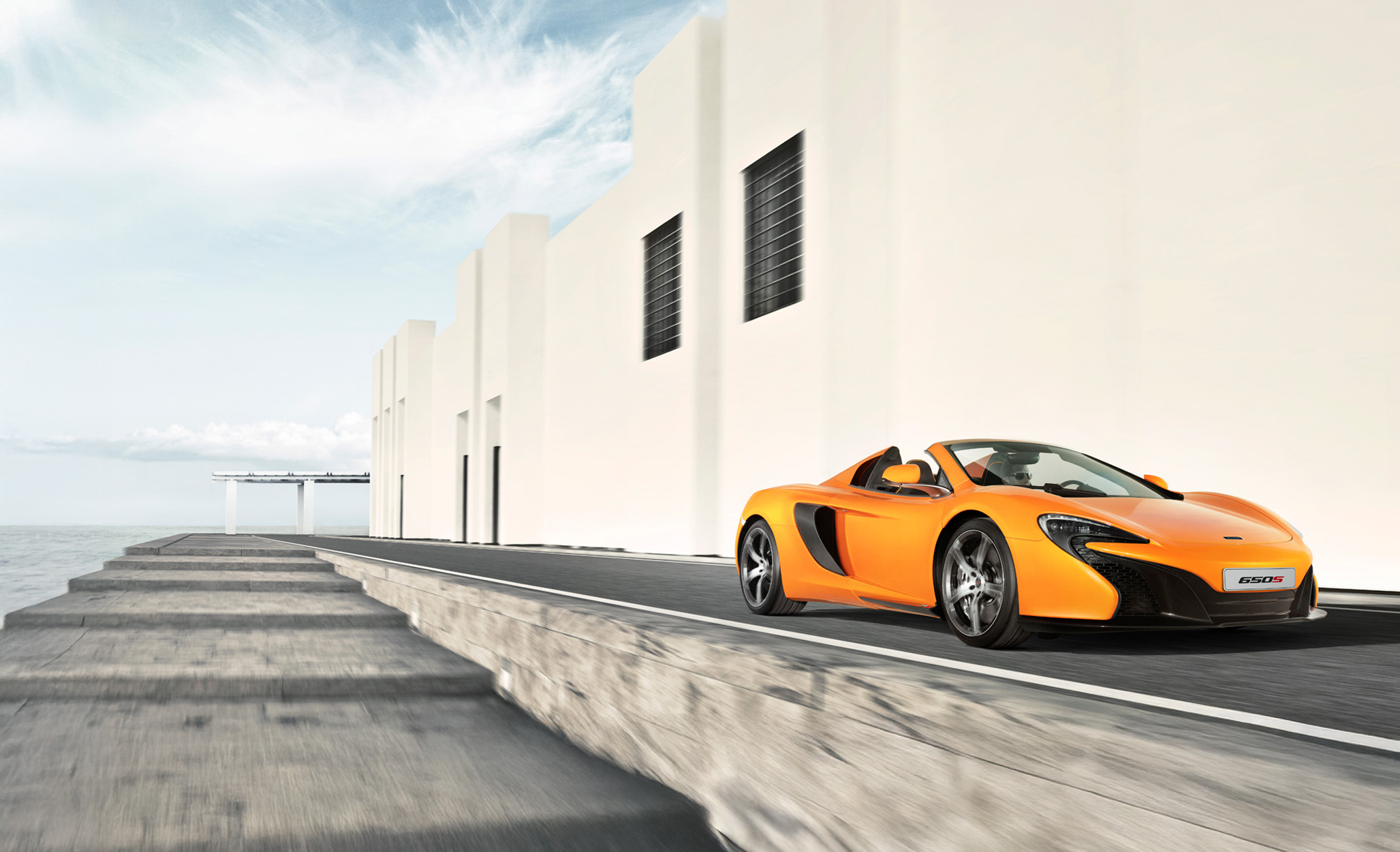 An orange McLaren 650S Spider driving down a road at speed, in the sun.