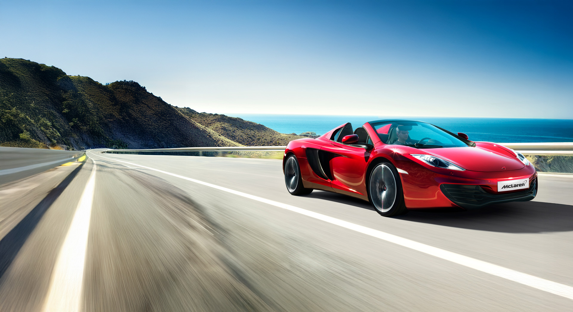Bright red McLaren 12C Spider driving along a coastal road at speed.