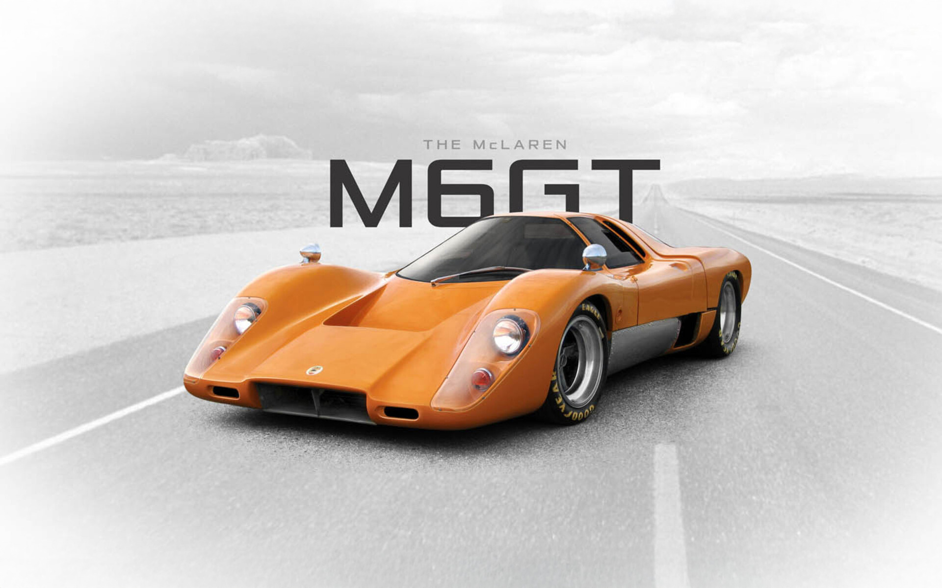 An orange McLaren M6GT parked on a remote road. The car is in colour while the background is faded black and white. Large text reading 'THE McLAREN M6GT' sits above/behind the car. 
