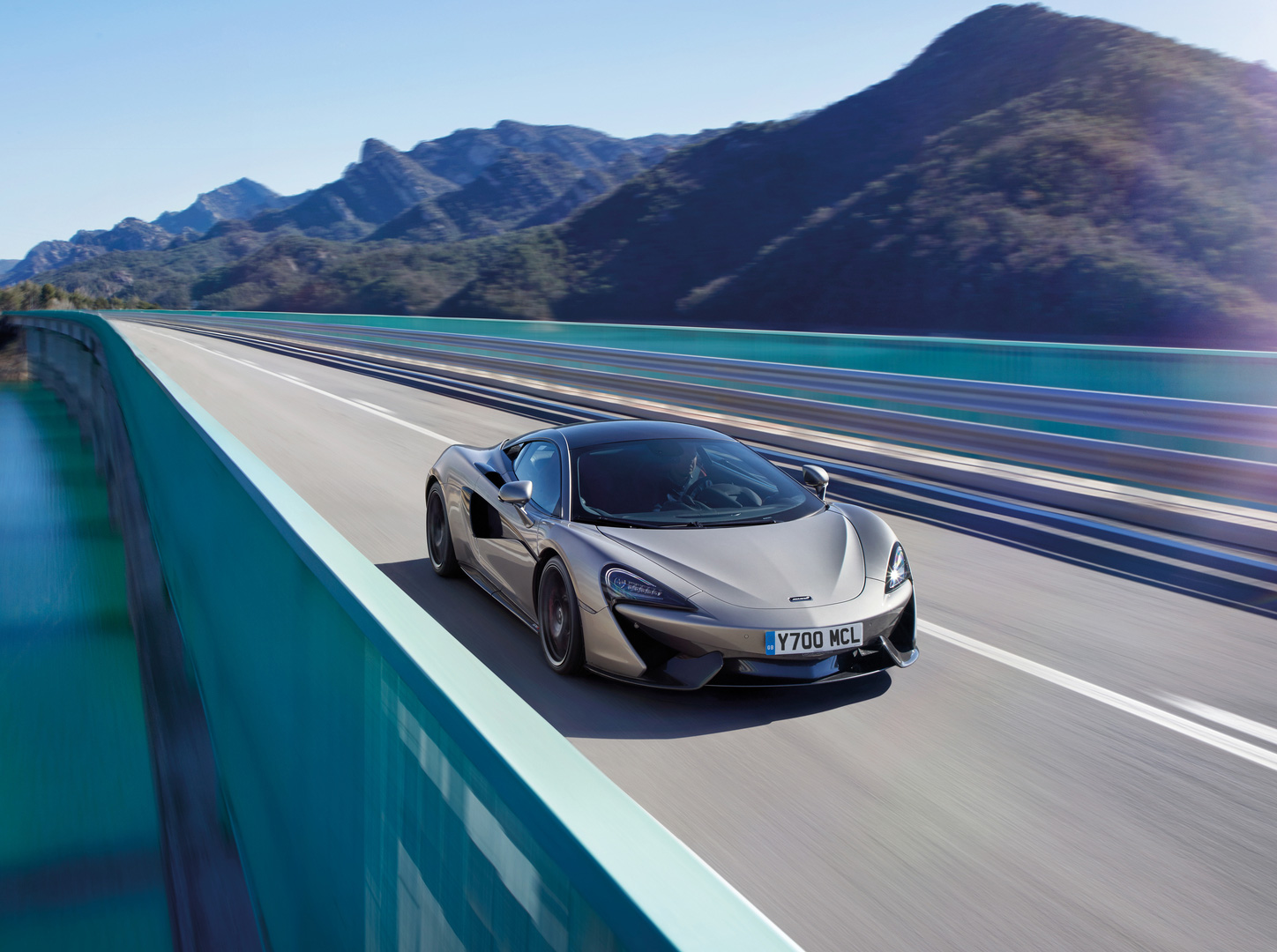 A bronze McLaren 570GT supercar driving across a bridge, with mountains and water in the background.