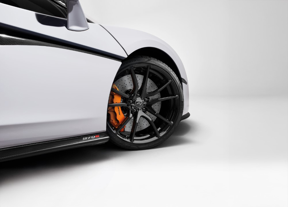 A close-up of the front wheel of a white McLaren 570S, showing the black alloy and orange brake caliper.