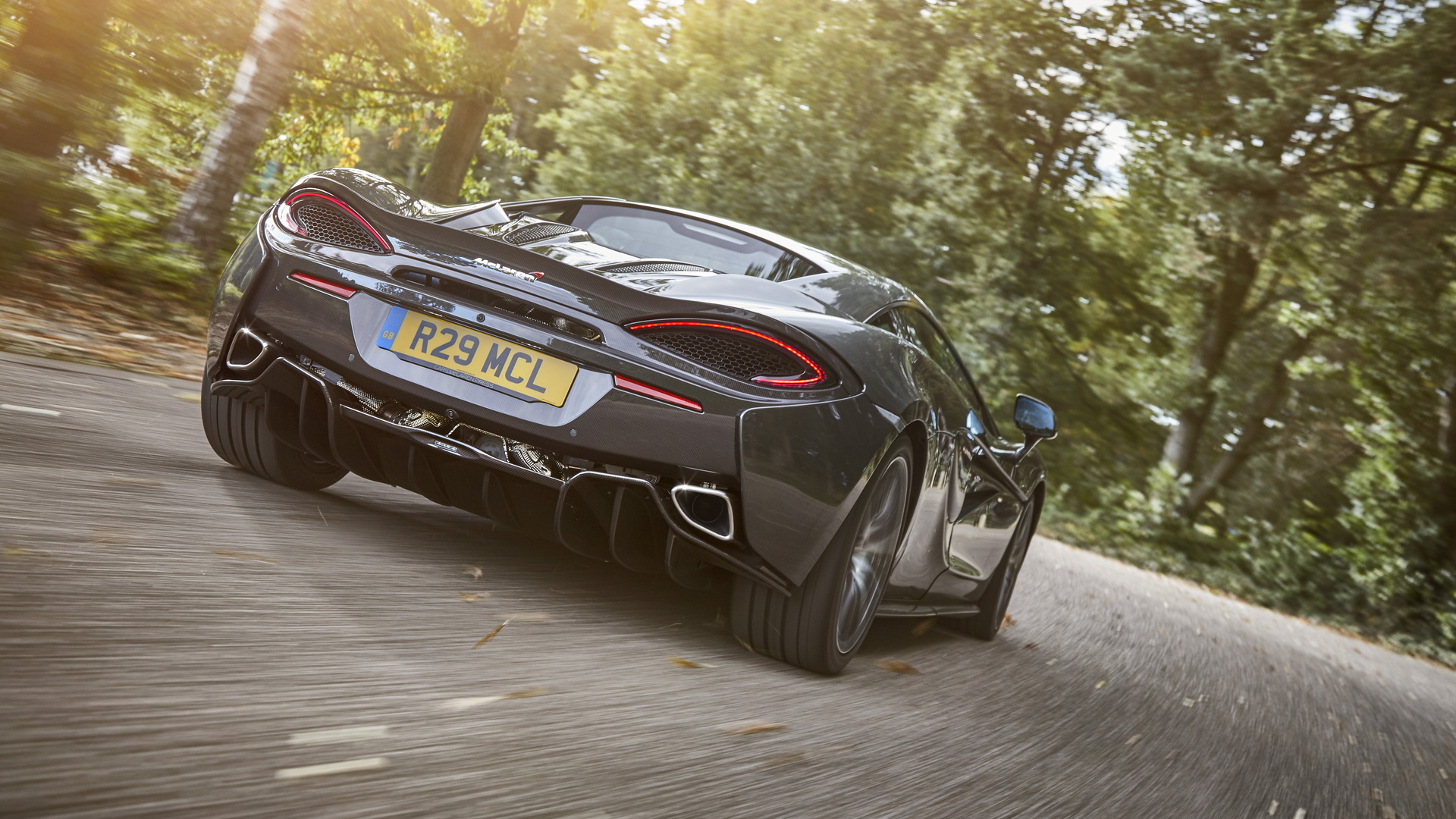 A rear-view action shot of a dark grey McLaren sports car driving away on a winding road. The image highlights the car's taillights, diffuser, and exhaust pipes.