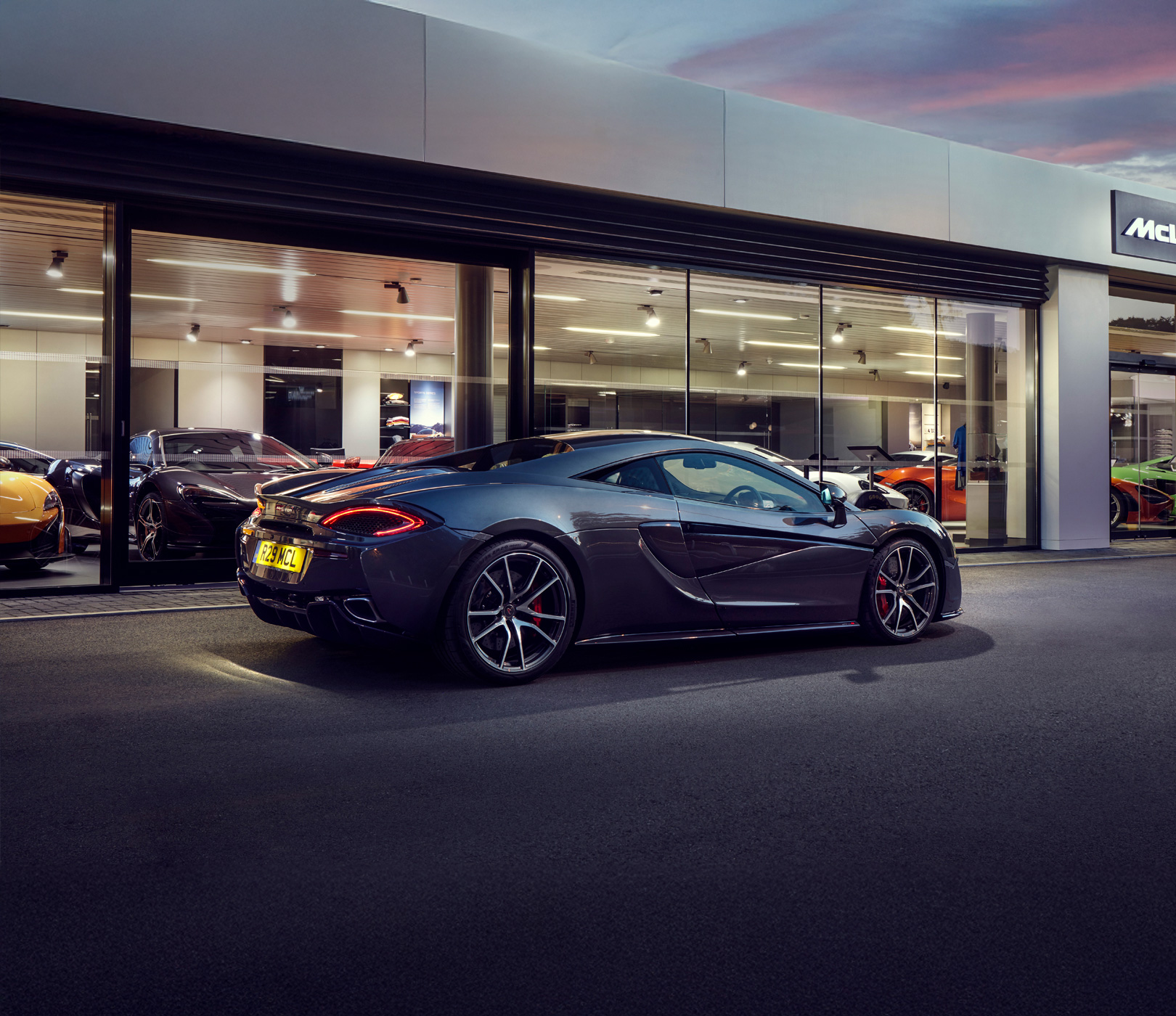 A grey McLaren 570S supercar parked outside a McLaren dealership showroom with large glass windows.