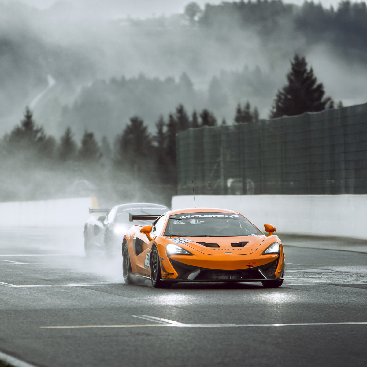 An orange McLaren 570S GT4 driving down the straight of a wet track, with spray coming off behind it.