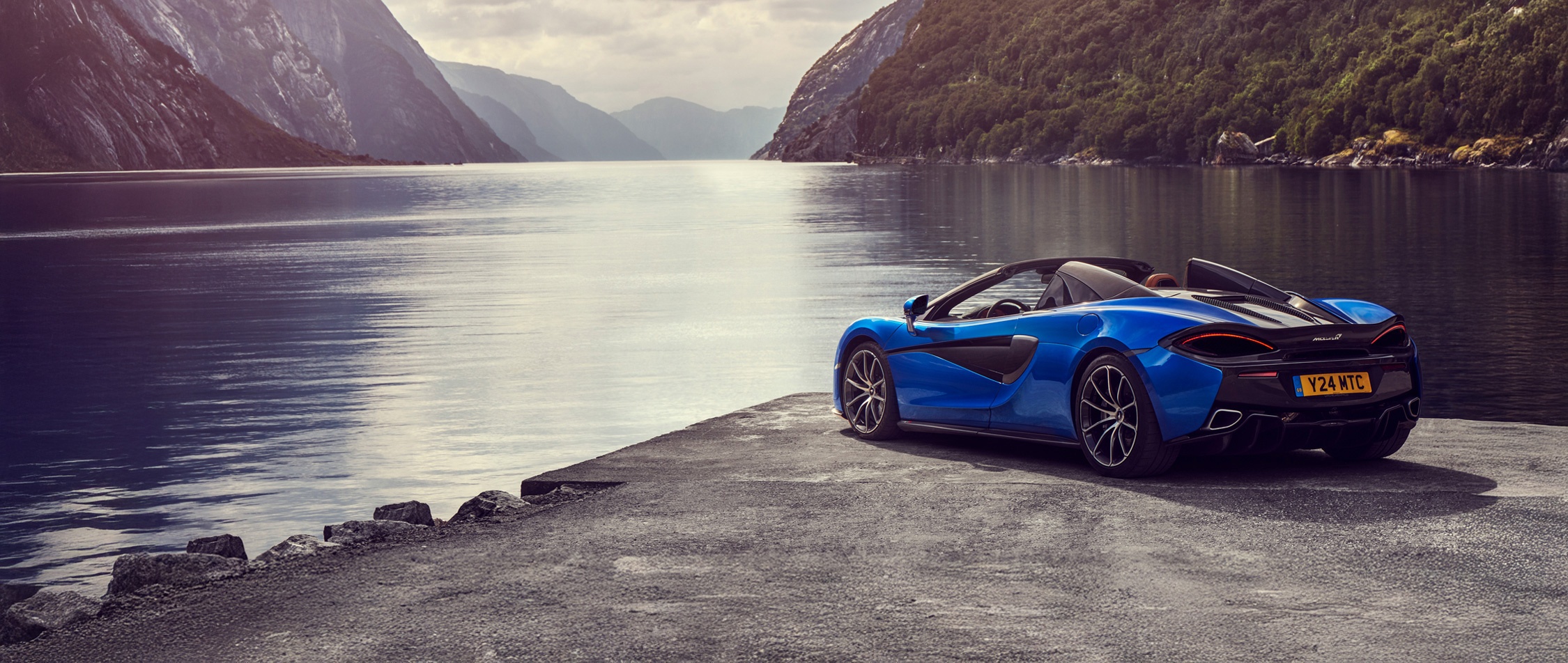 A blue McLaren 570S Spider convertible is parked by a scenic lake with a large mountain or cliffside in the background, showcasing its side profile