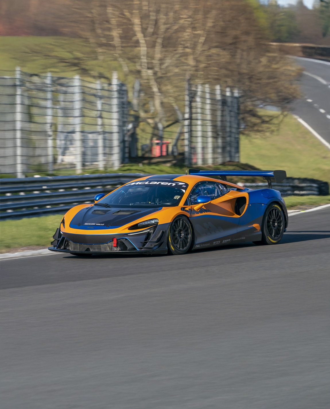 An orange, blue and black McLaren Artura GT4 driving around a race track.