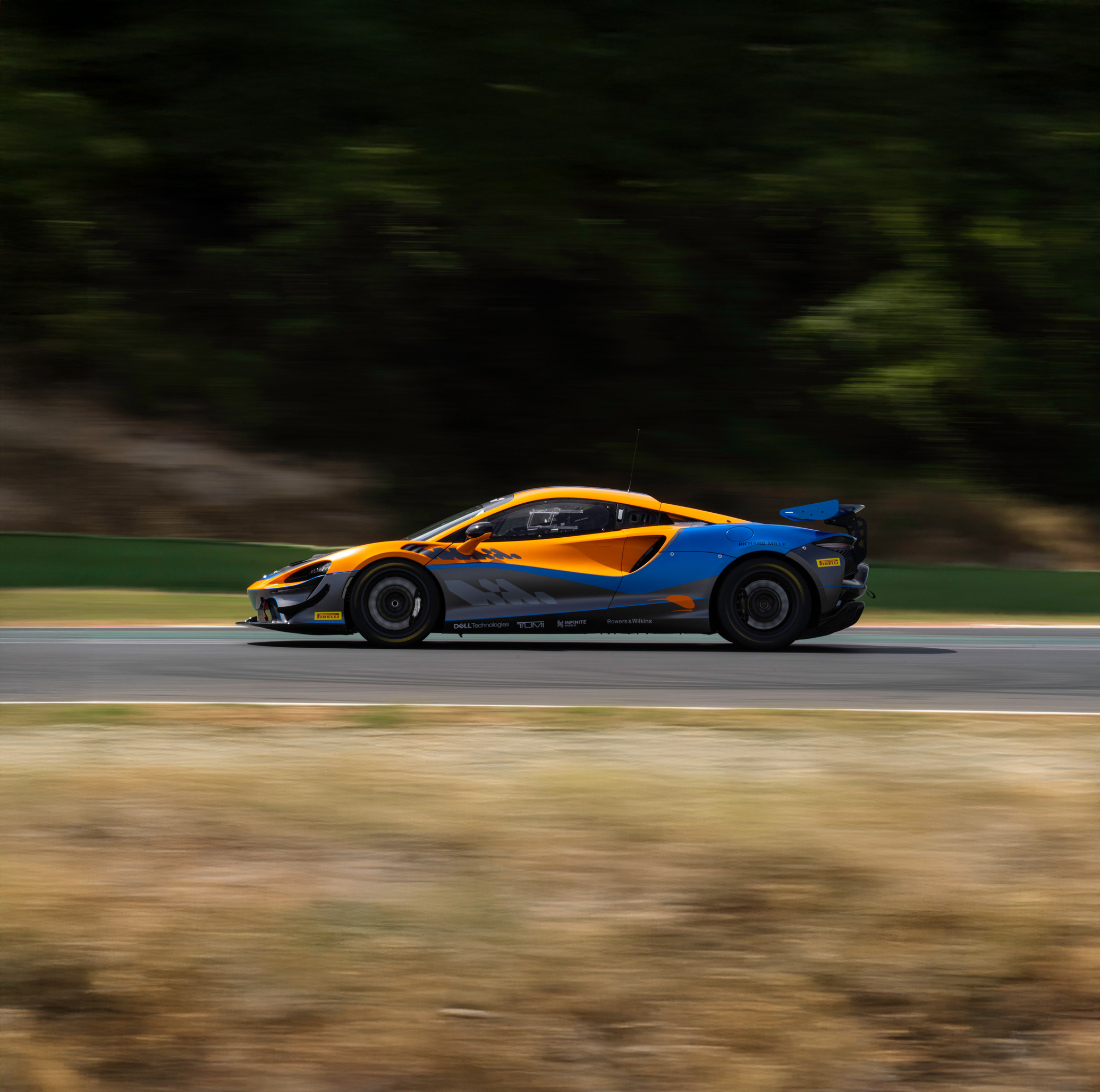 Side view of an orange, blue and black McLaren Artura GT4 driving around a race track at speed.