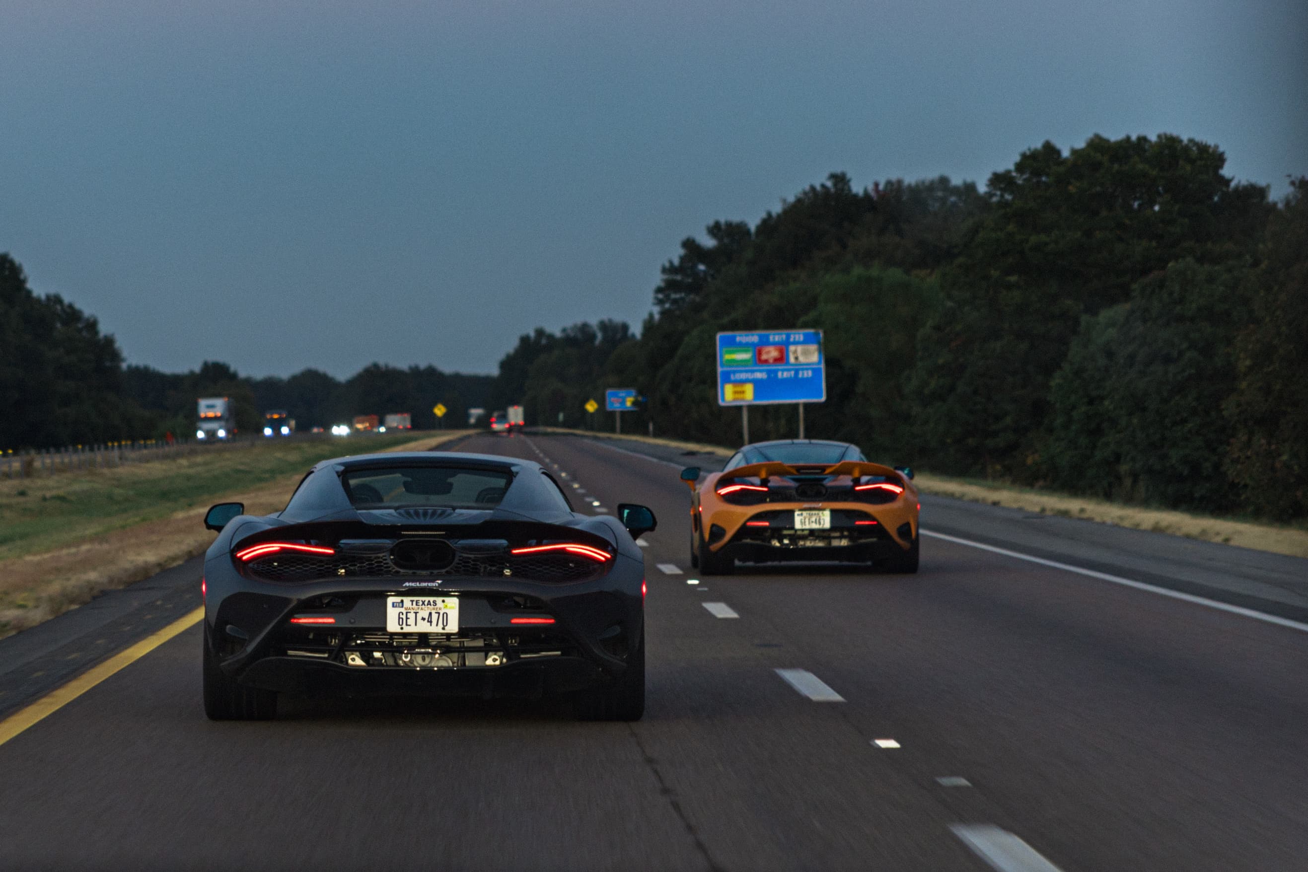 A black McLaren and an Orange McLaren driving in two lanes next to each other at dusk. A scene from Day 8 of the 'McLaren States of Endurance' road trip. 