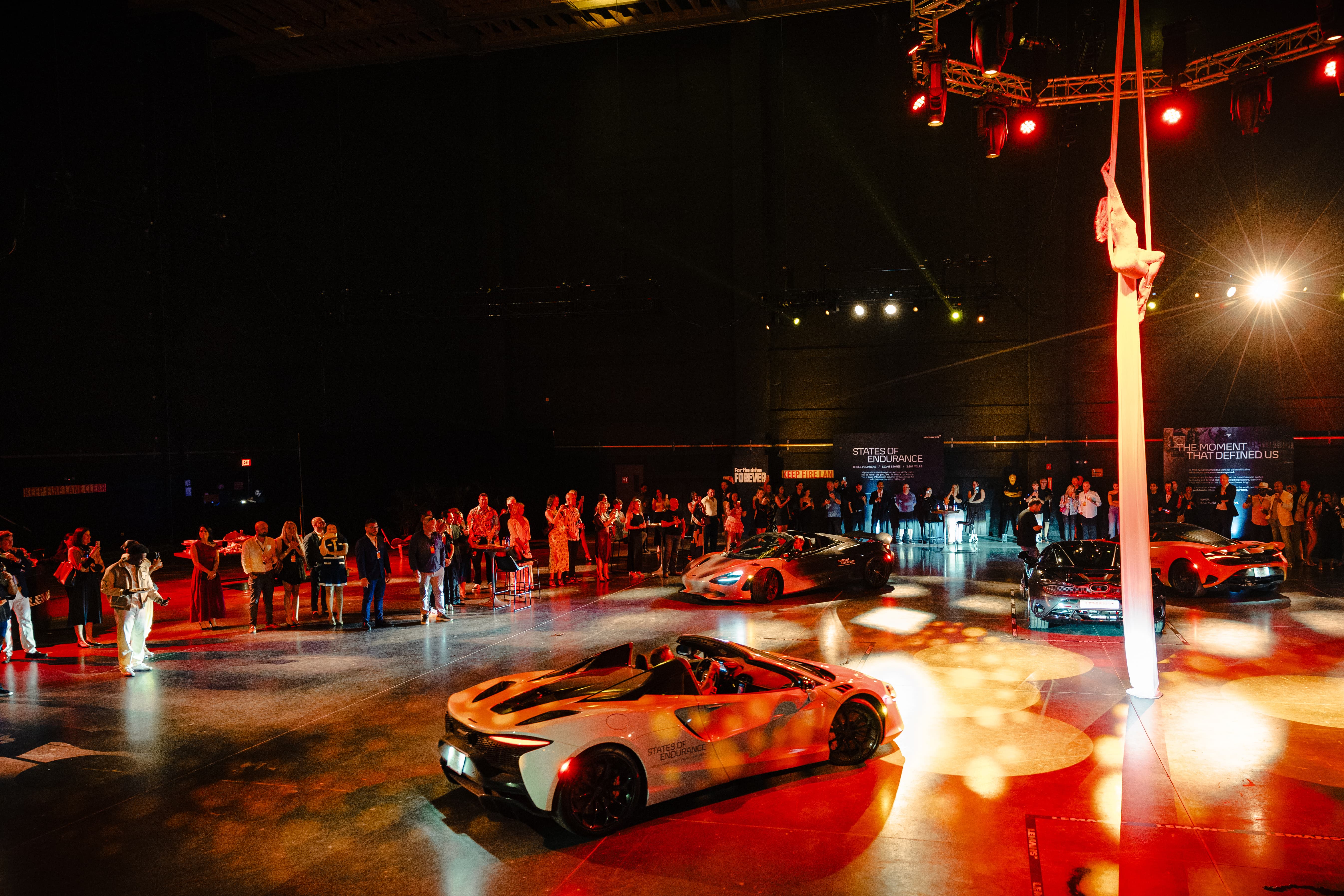 Crowds looking at the McLaren States of Endurance cars parked in an exhibition setting under theatrical red and orange spotlights. A scene from Day 9 of the 'McLaren States of Endurance' road trip. 