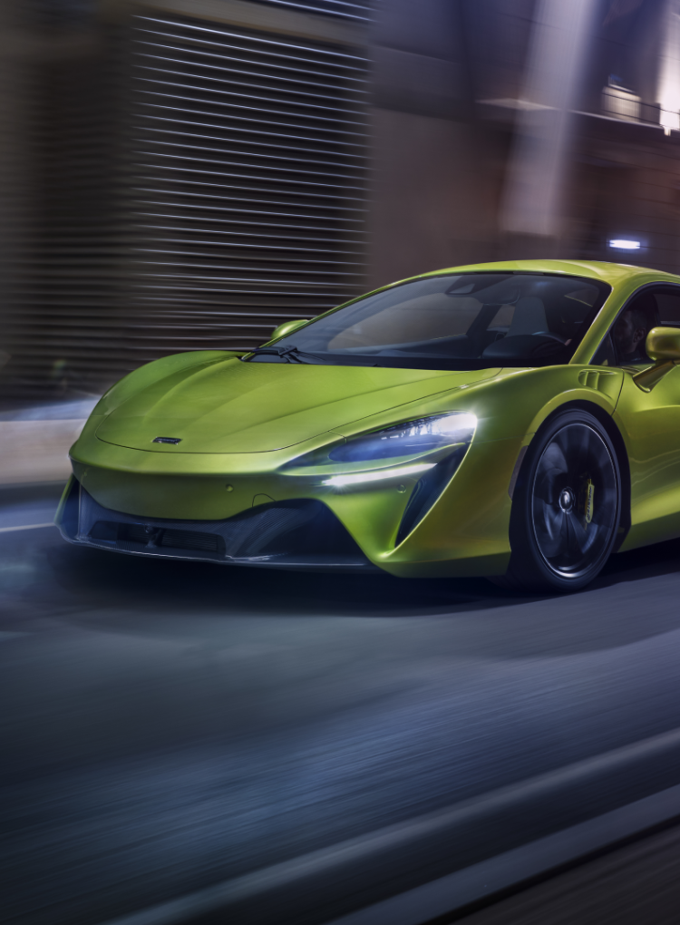 The McLaren Artura hybrid supercar in a vibrant yellow-green, driving through an urban setting with a motion blur effect.