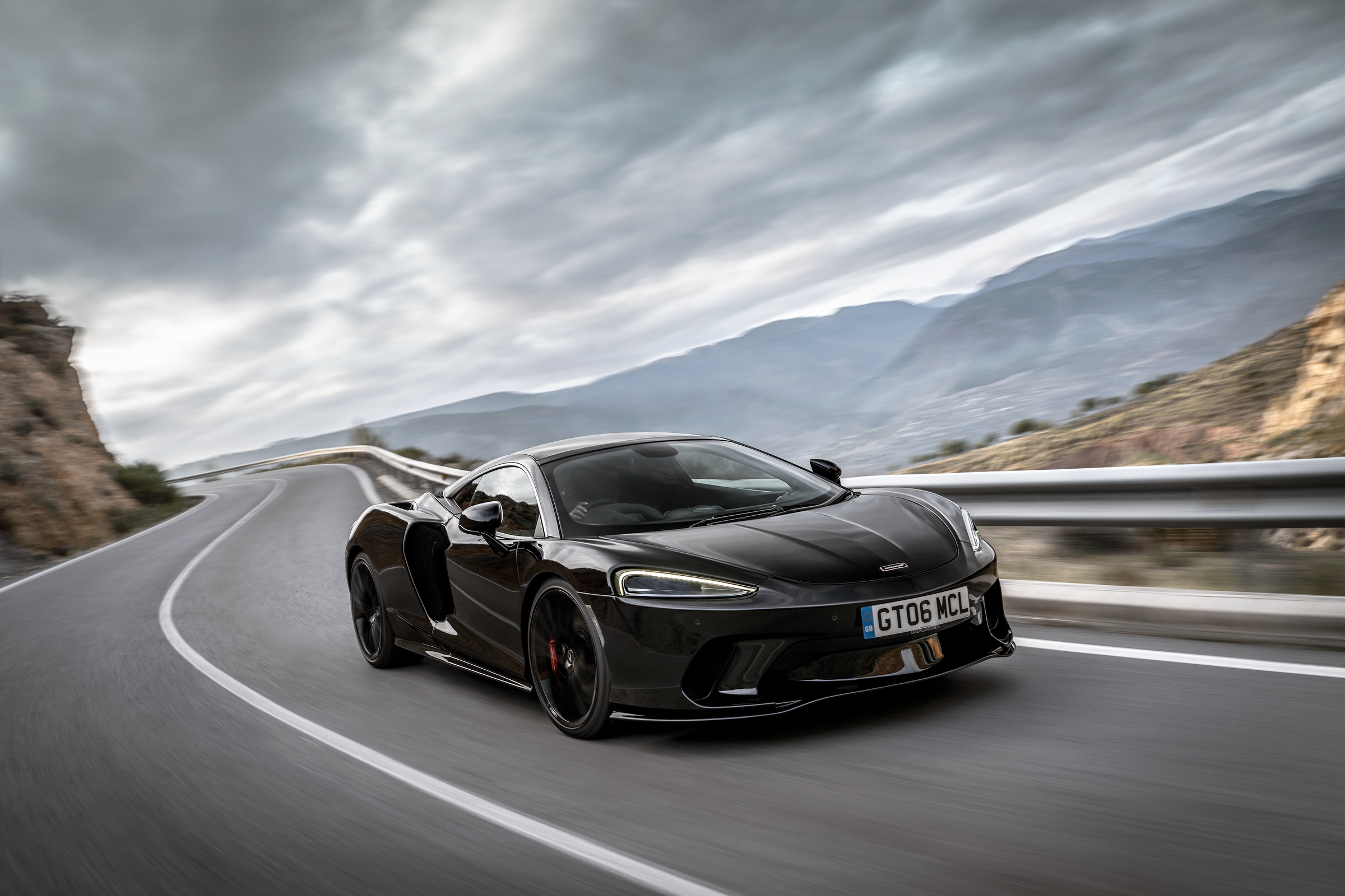 The black McLaren GT in a dynamic motion shot, driving on a road with mountains and a cloudy sky in the background.
