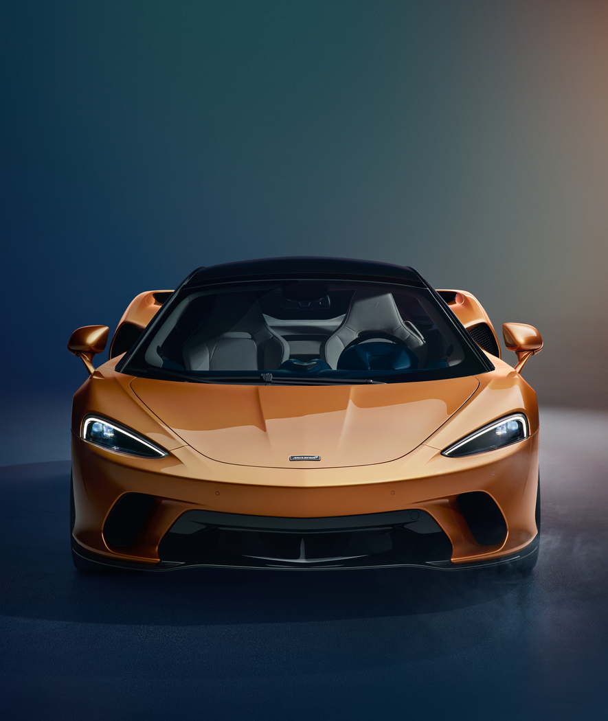 A front-on view of the McLaren GT in a bespoke gold finish, with its headlights on, in a dark studio setting.