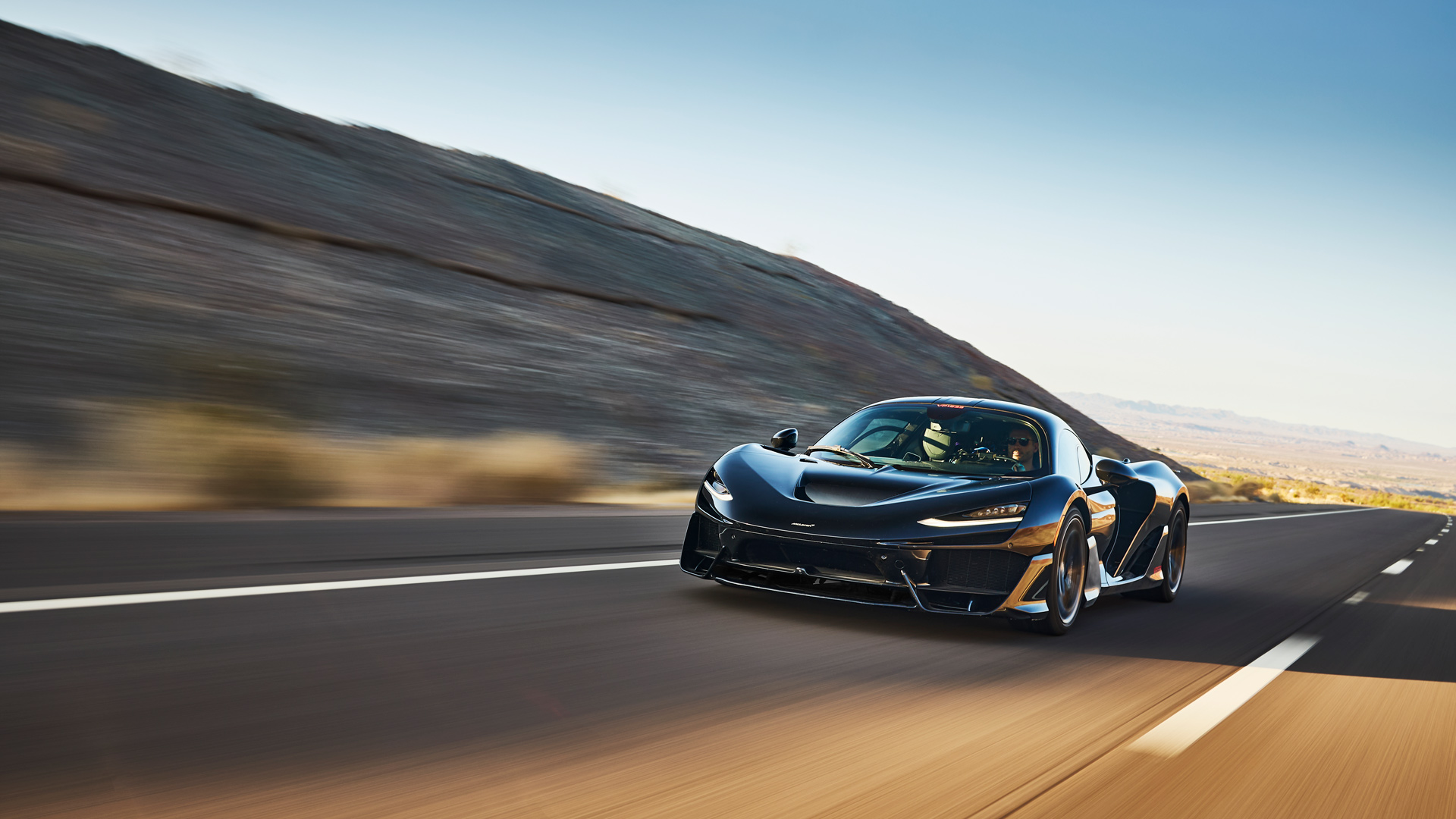 Dynamic front shot of the black McLaren W1 prototype on a winding desert road. The hypercar's powerful presence and Formula 1 aerodynamics are visible, showcasing the W1's road manners during the final phase of extreme heat testing.