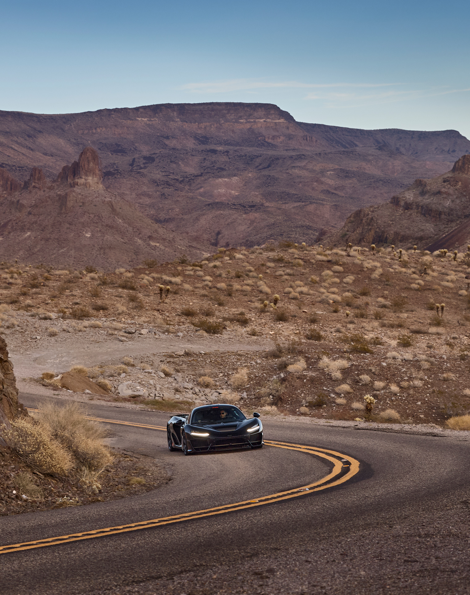 Black McLaren W1 prototype driving on a winding desert mountain road amidst rugged terrain. Front-on shot emphasizes the extreme durability testing in hot, arid conditions, relevant for McLaren performance and Ultimate Series off-track evaluation.