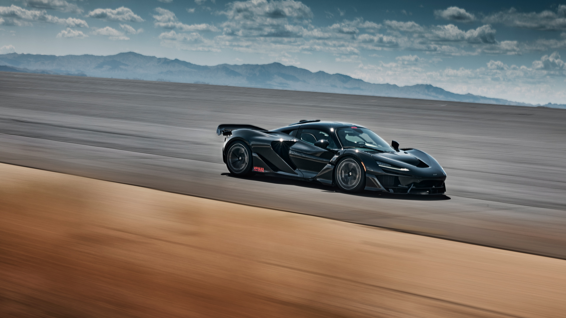McLaren W1 Ultimate Series hypercar prototype performing high-speed dynamics tests on a wide desert track. Black test mule showcases aggressive active aerodynamics and Formula 1-inspired downforce during hot climate validation.