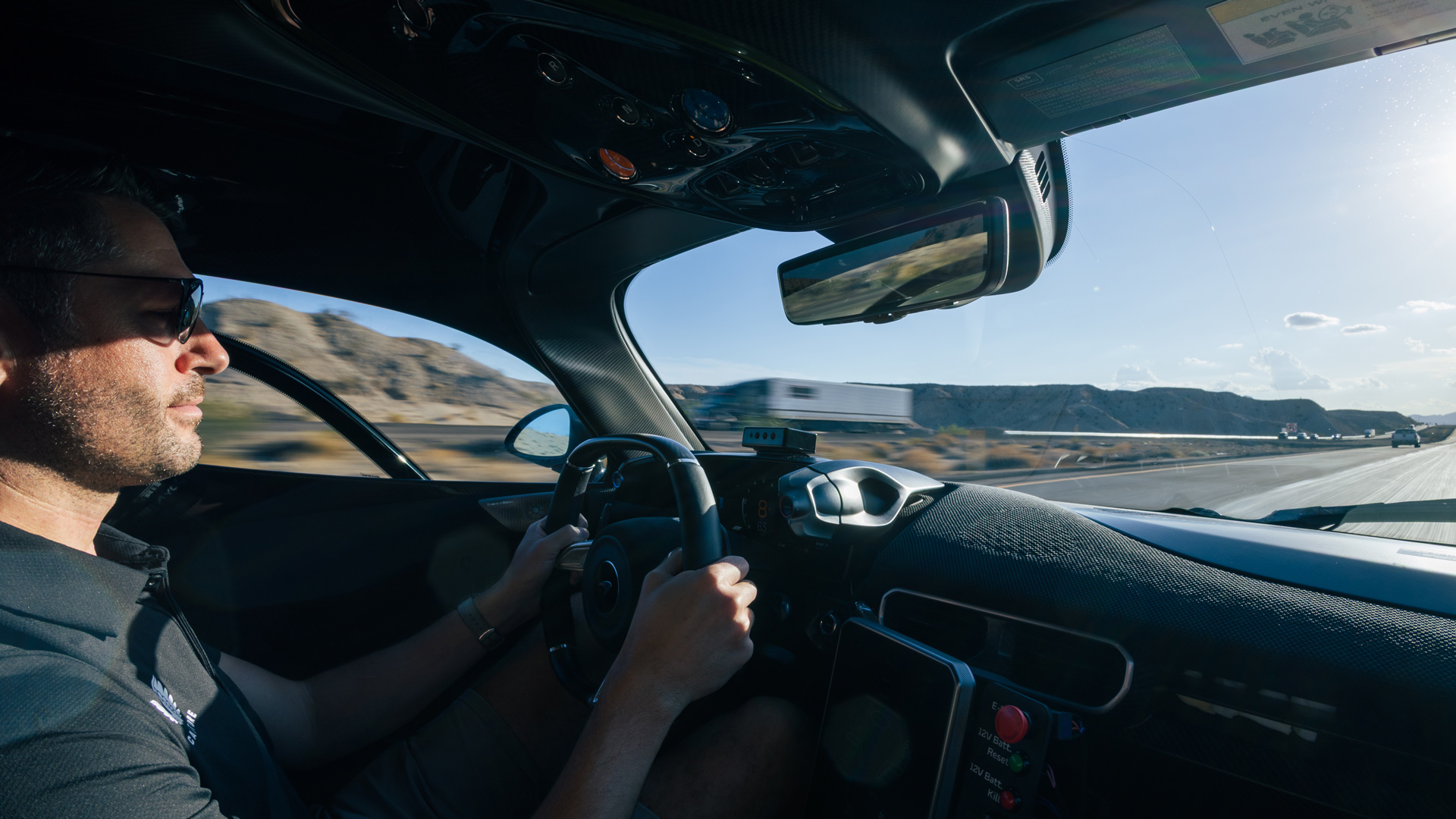 In-car POV of a test driver wearing sunglasses, intensely driving the McLaren W1 hypercar prototype down a desert road. Highlights the supercar cockpit, steering wheel, and extreme conditions of the McLaren hot weather validation.