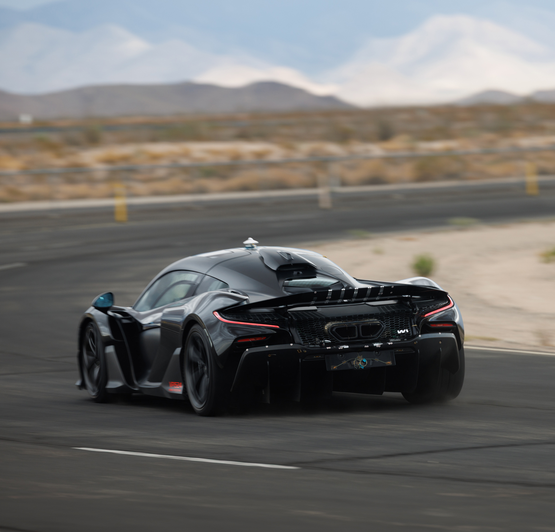 Rear view of the black McLaren W1 hypercar prototype cornering aggressively on a test track in a hot, arid environment. The image emphasizes the huge active rear wing, carbon fibre diffuser, and twin high-mounted exhausts of the 1275PS Ultimate Series car during limits testing.