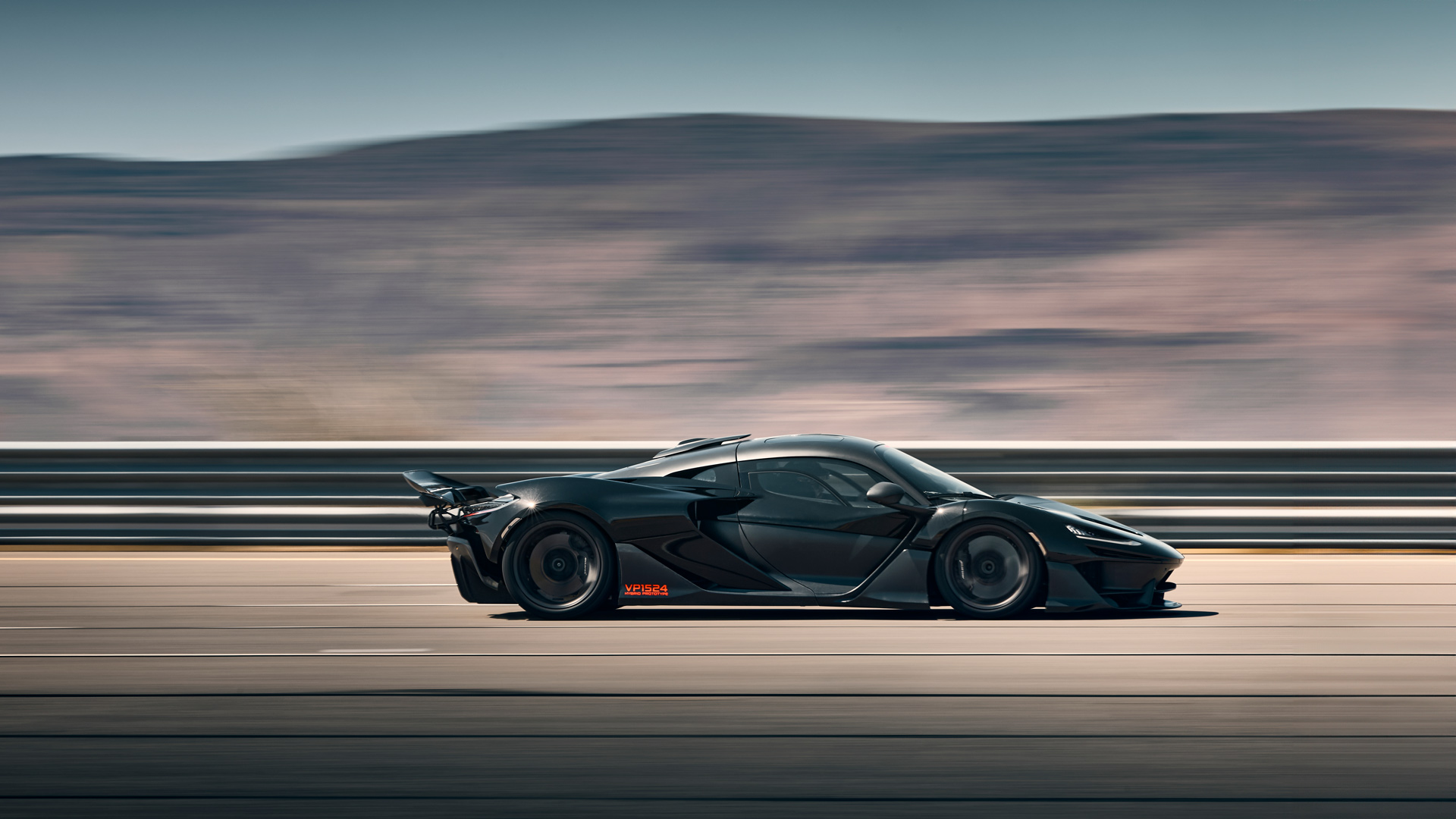 Side profile motion shot of a black McLaren W1 prototype accelerating on a test track or closed highway. The image shows the low-slung body, aggressive side vents, and large rear wing, highlighting aerodynamic testing and high-speed handling.