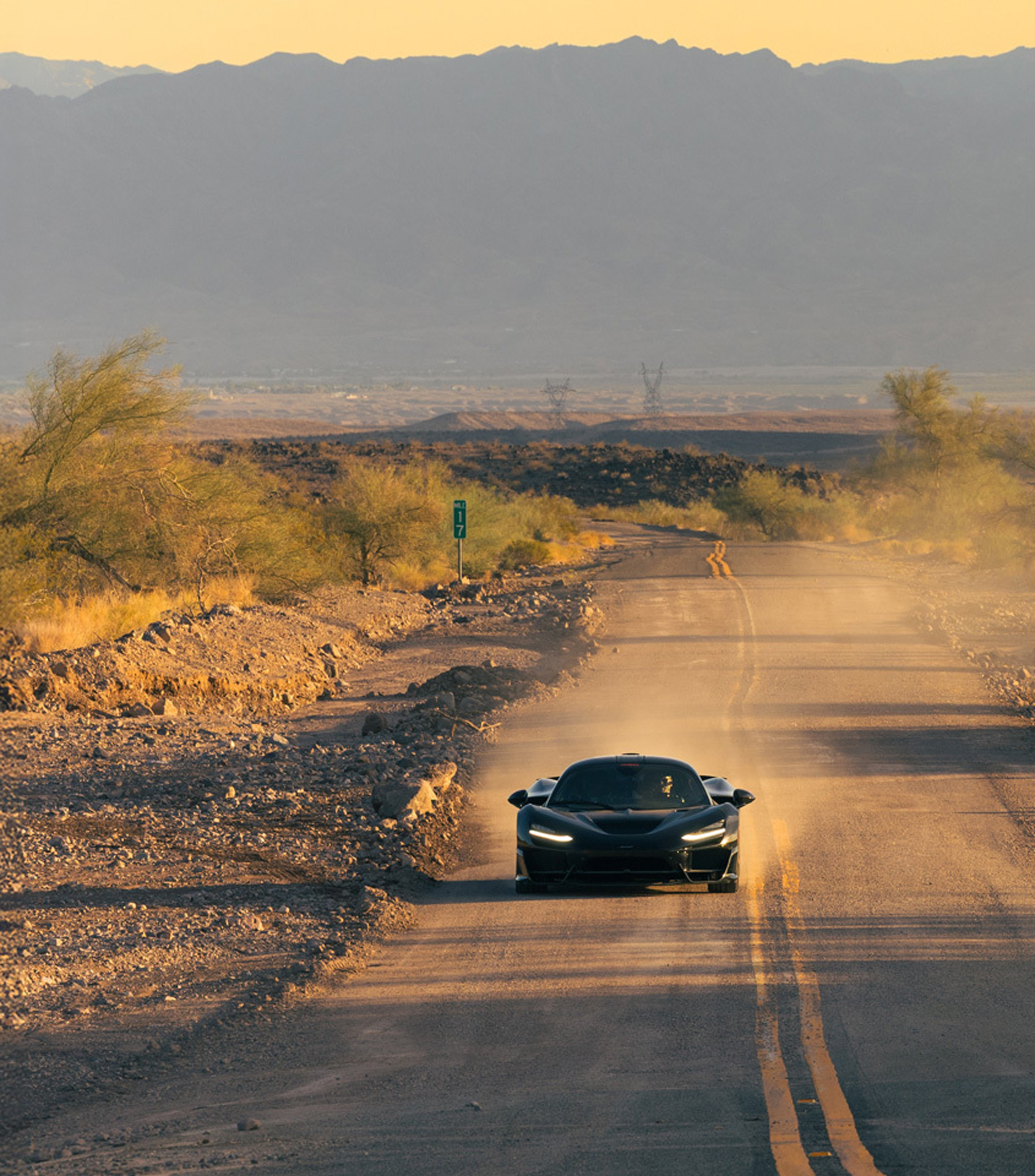 McLaren W1 prototype kicking up dust on a long, remote desert road at sunset. Front view of the Ultimate Series supercar in black during rigorous hot climate development testing for durability and cooling performance.
