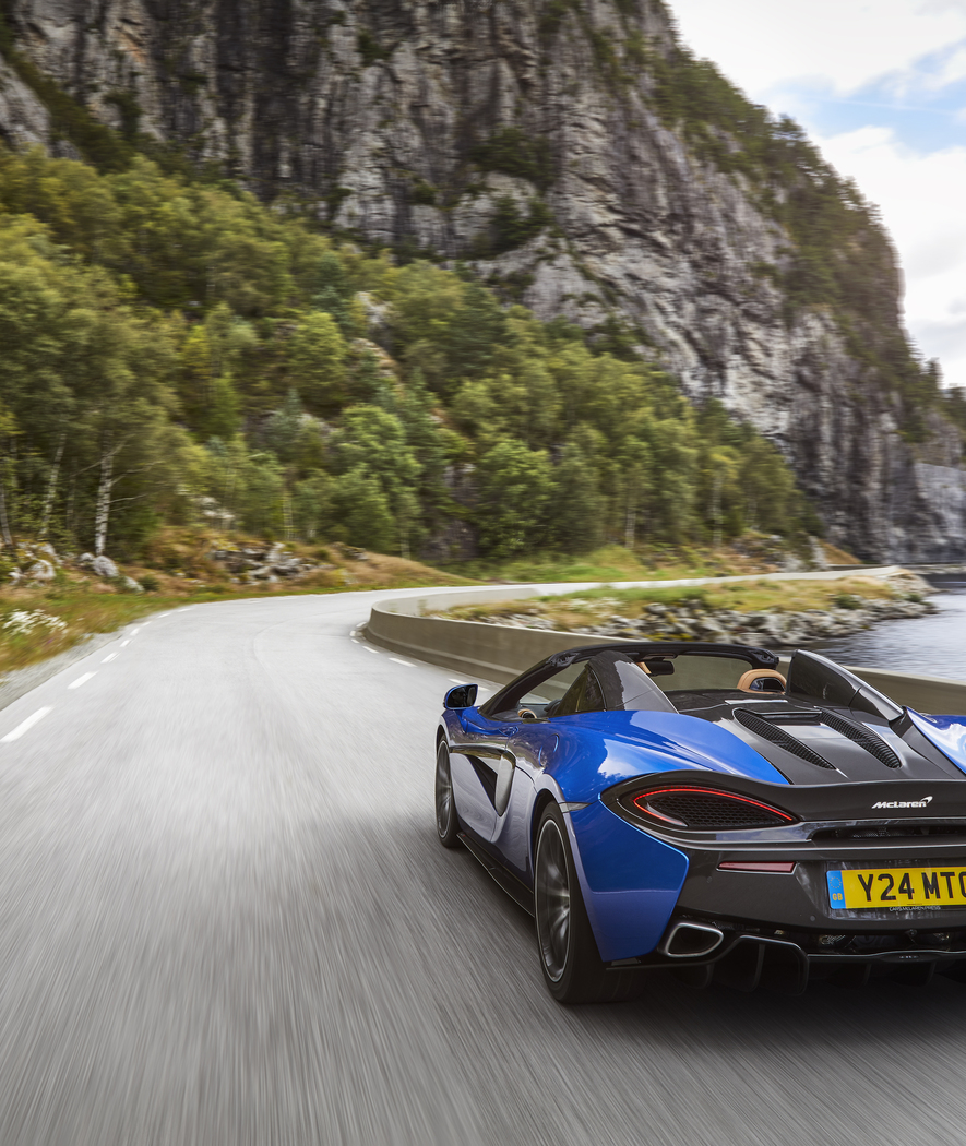 The rear of a blue McLaren 570S Spider, a convertible, driving on a scenic road along a body of water.