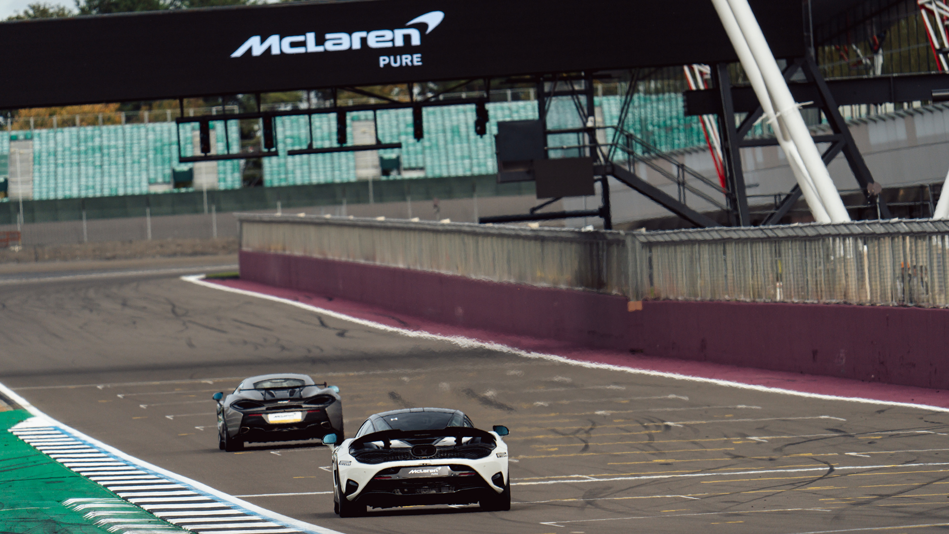 A rear view shot of the McLaren 750S and 620S driving down the Silverstone track with a McLaren sign across the track.