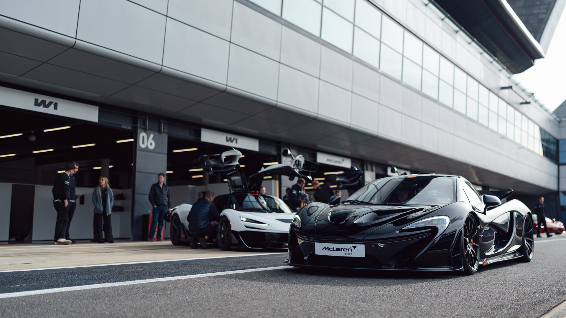 A black McLaren W1 parked on within the pit lane of a racetrack with a white McLaren W1 stationary in the background. 