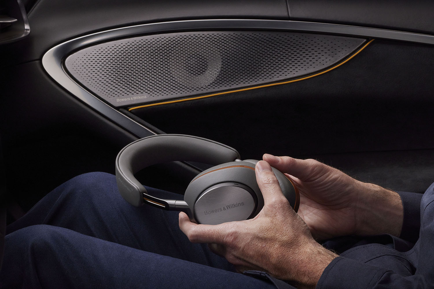 Image showcasing the Bowers & Wilkins speakers as part of the McLaren car interior, as well as a man sat in the car, holding the PX8 McLaren Edition headphones.