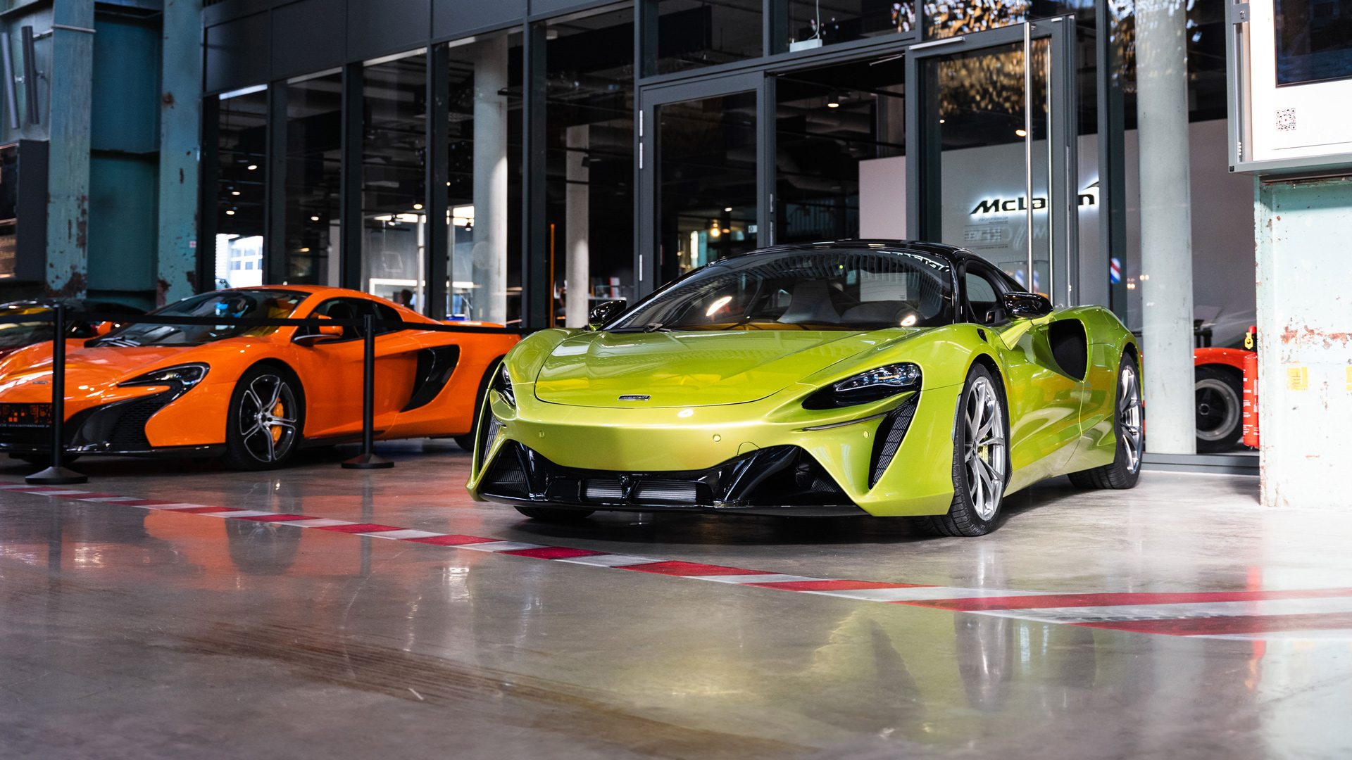 McLaren Munich official retailer showroom featuring two supercars: A Flux Green McLaren Artura Hybrid in the foreground and a McLaren Orange Artura Hybrid in the background.