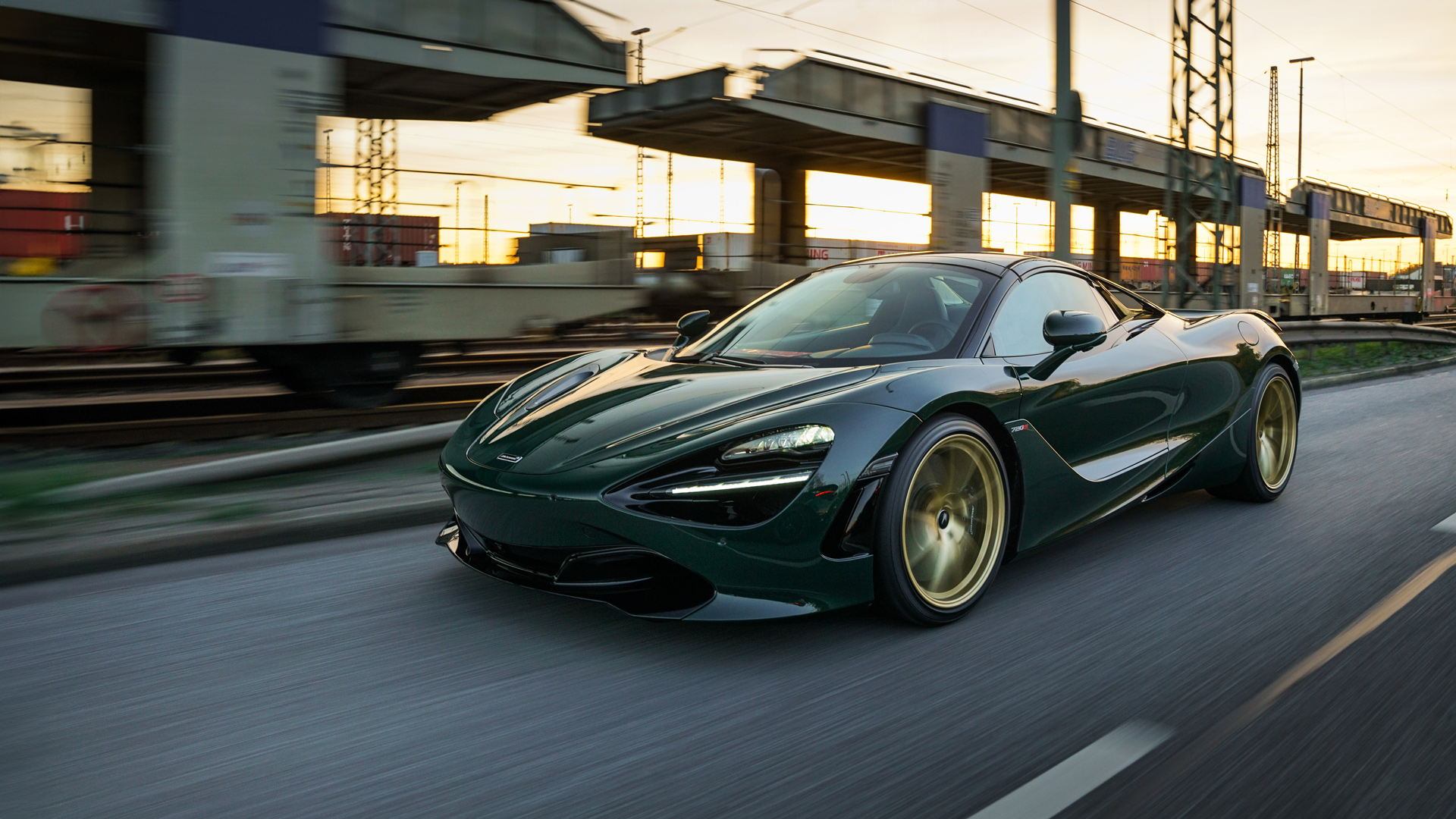 McLaren 750S supercar in a bespoke British Racing Green (or MSO Green) paint finish, moving dynamically alongside a moving train in an urban/industrial setting near Hamburg, Germany. The high-performance coupe features custom gold alloy forged wheels and a carbon fibre aerodynamic kit.