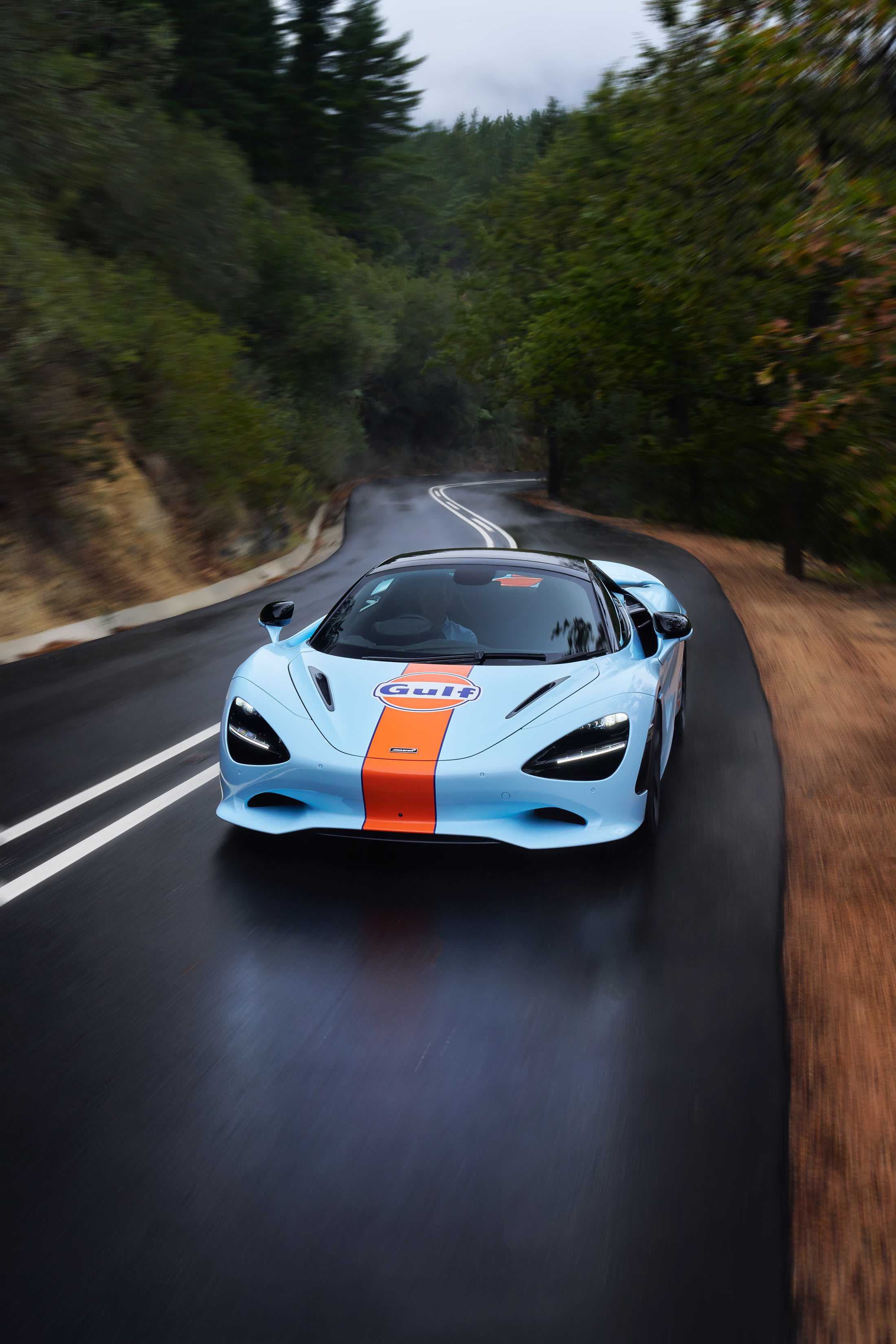 A Gulf branded McLaren car driving on a wet forrest road. 