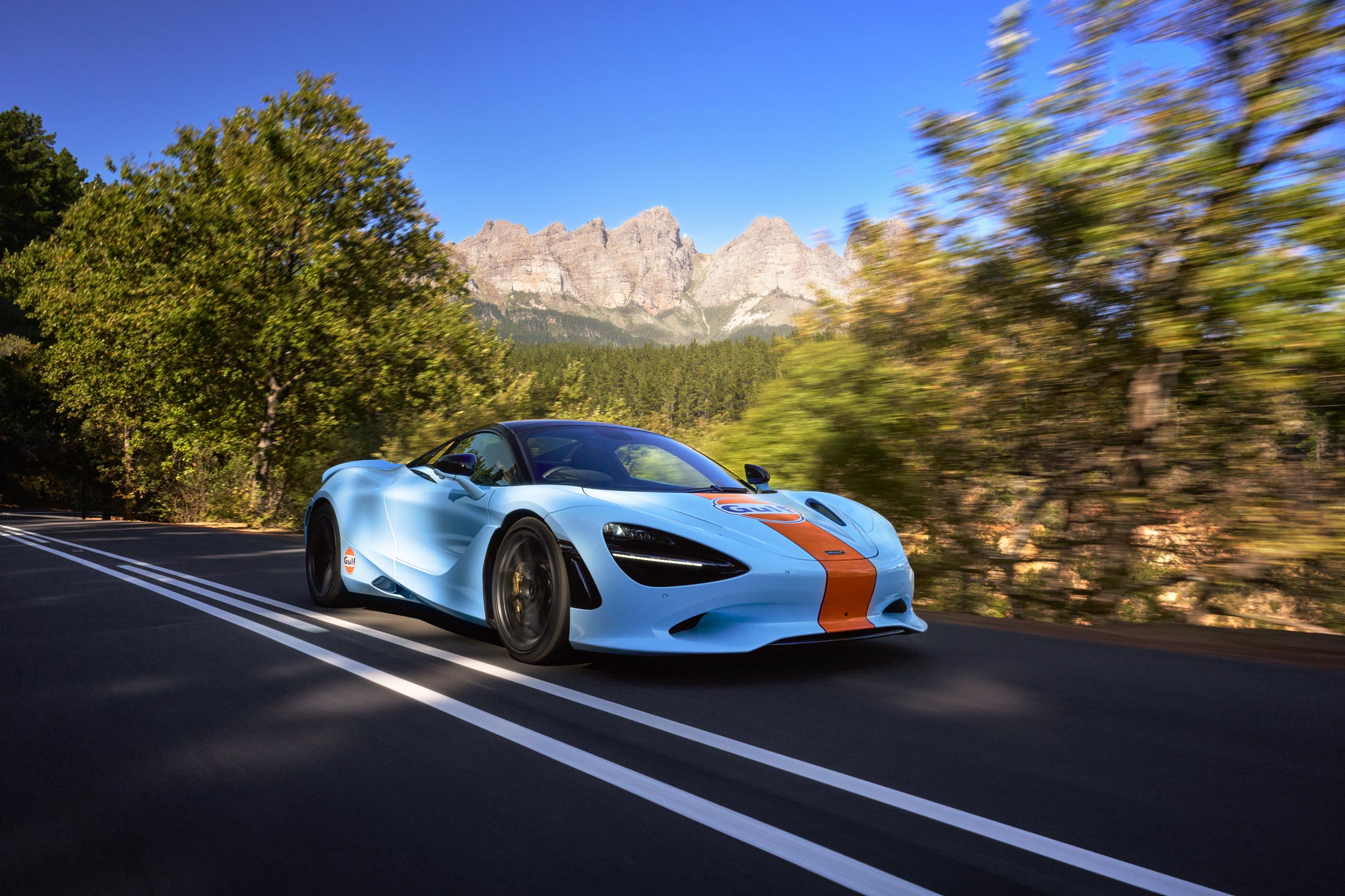 Gulf Branded McLaren driving down a road at speed, with mountains behind it.
