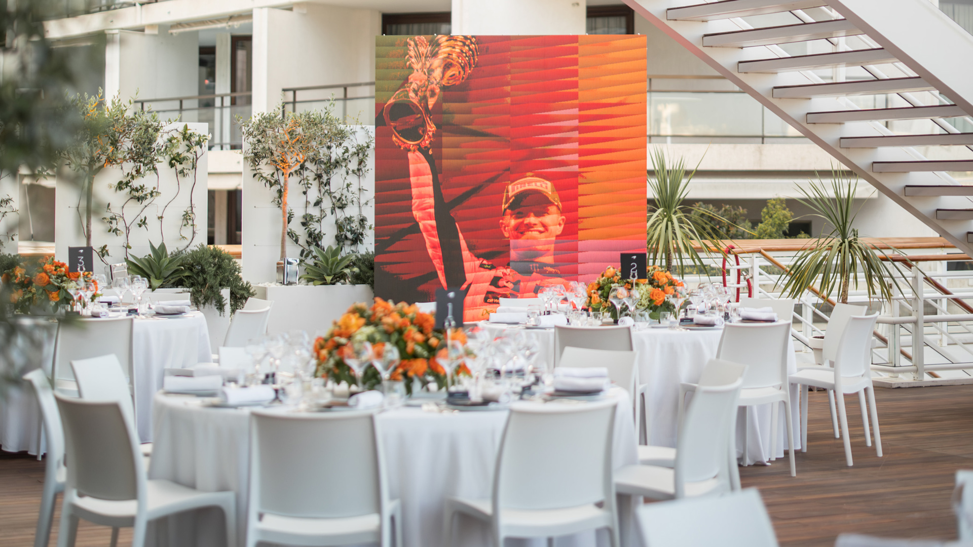 Round tables set for a sophisticated McLaren F1 hospitality dining event with a large digital screen displaying a McLaren driver celebrating a victory, emphasizing the exclusive race experience.