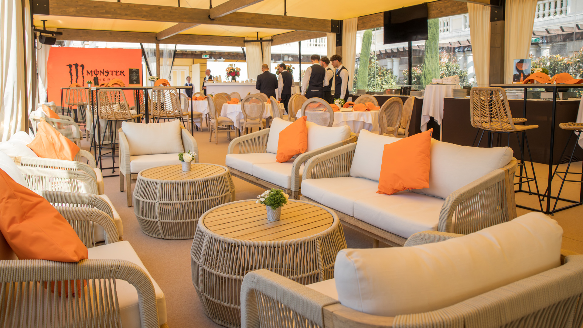 Luxurious outdoor lounge seating and dining area in the McLaren F1 hospitality suite at the Monaco Grand Prix, featuring orange pillows and Monster Energy branding.