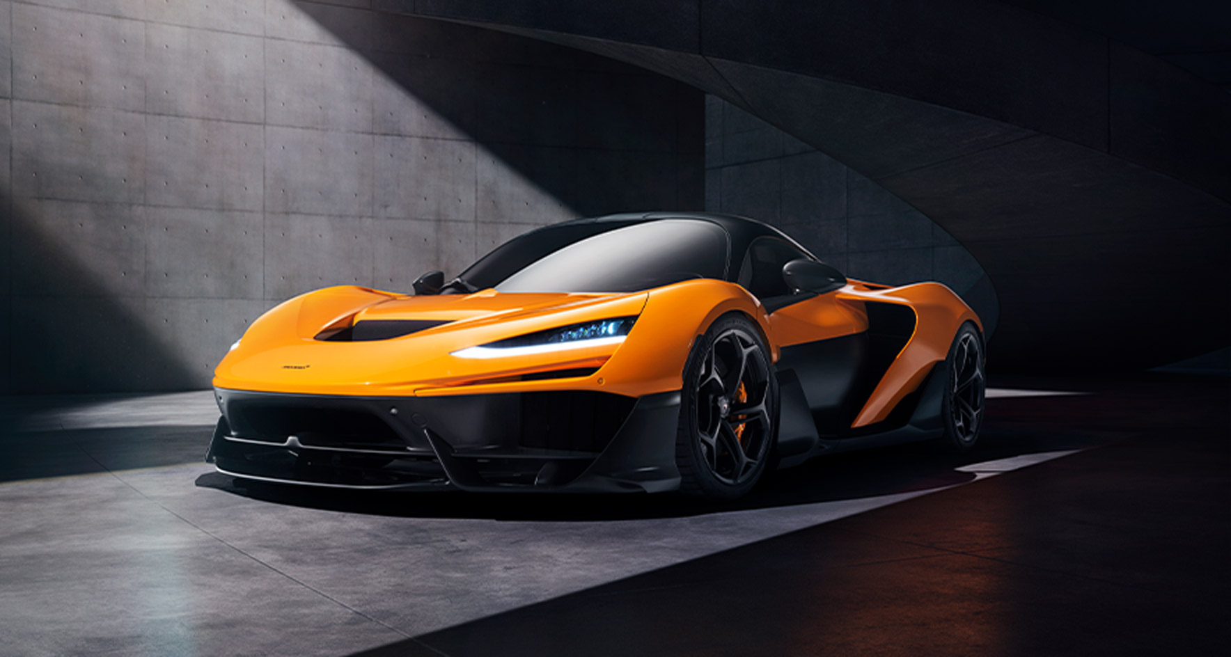 A front three-quarter view of the orange McLaren W1 parked in a modern, concrete garage, emphasizing its sleek headlights and aerodynamic front splitter.