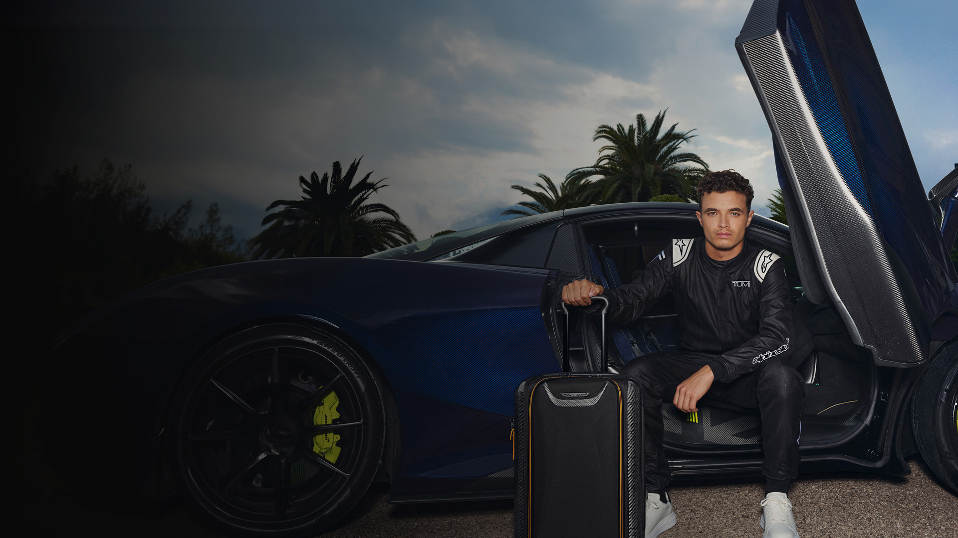 Lando Norris sitting in the open doorway of a blue McLaren holding a McLaren TUMI suitcase