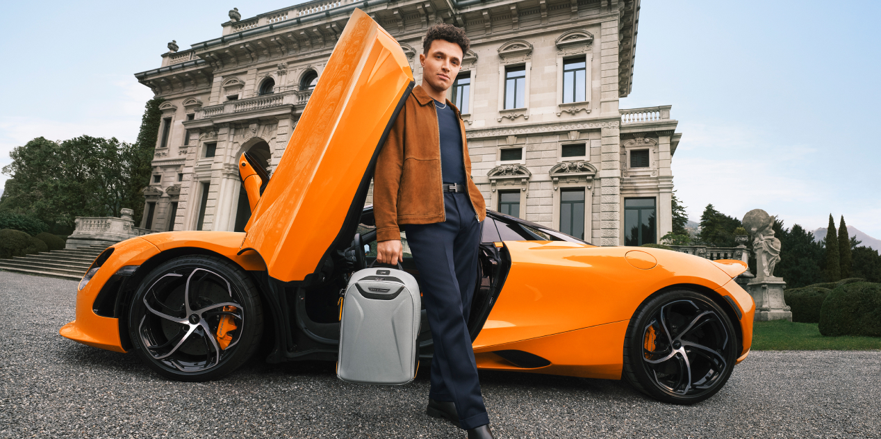 Lando Norris stood, leaning on the door of an orange McLaren holding a super grey McLaren TUMI backpack.