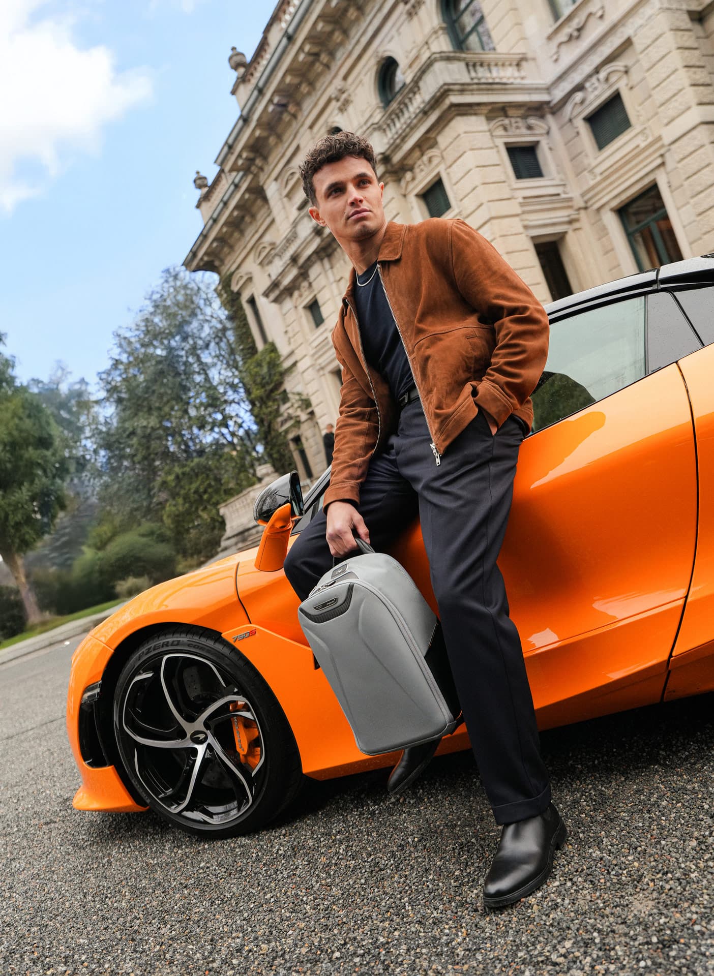 Lando Norris stood, leaning on the door of an orange McLaren holding a super grey McLaren TUMI backpack.