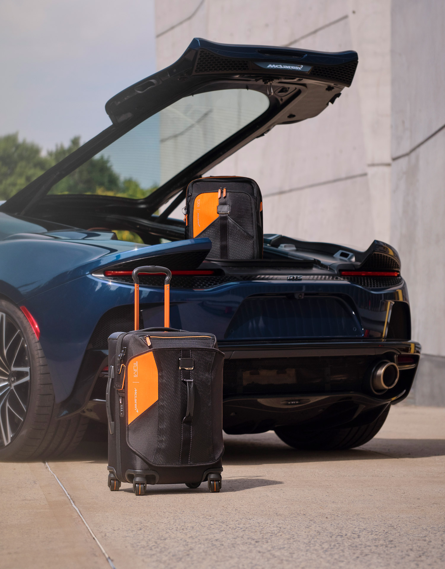Two black and orange TUMI | McLaren bags sat by the rear of a blue McLaren GTS, with its trunk open.