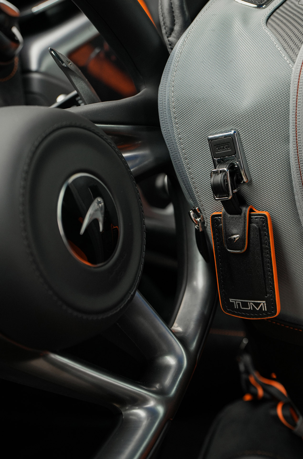 Close up of Tumi | McLaren luggage tag hanging from a grey backpack, next to a McLaren steering wheel.