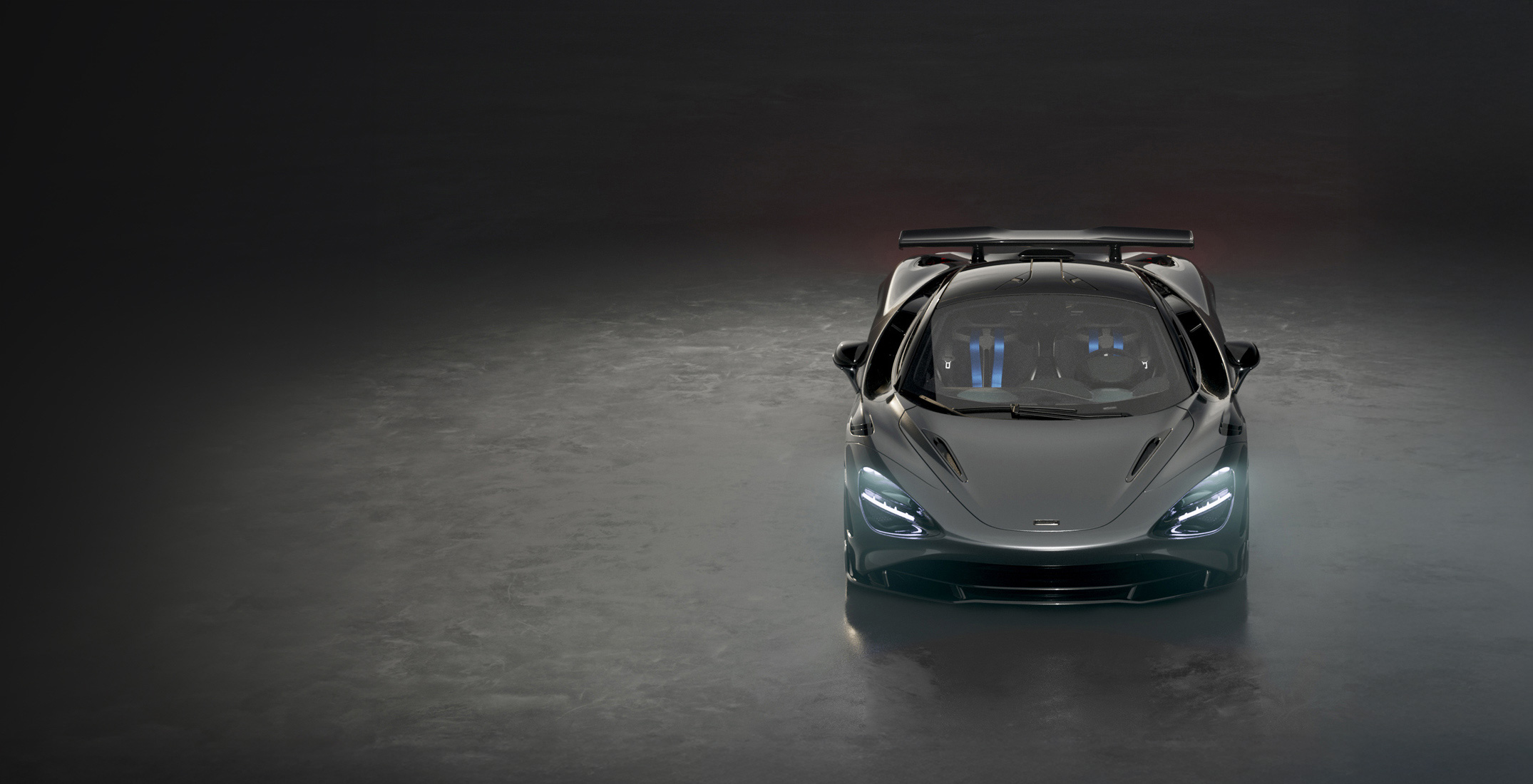 A dark grey McLaren 750S Le Mans special edition supercar is shown from a front-on view in a dark studio, with its headlights on and a prominent rear spoiler.