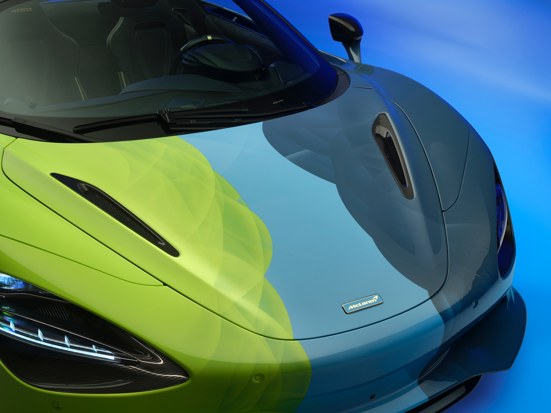 The nose of the Project Chromology 750s by MSO and Nat Bowen, on display against a glowing blue background. The car is various overlaying shades of green and blue, here you can see details of the overlapping paint and the blue McLaren badge.