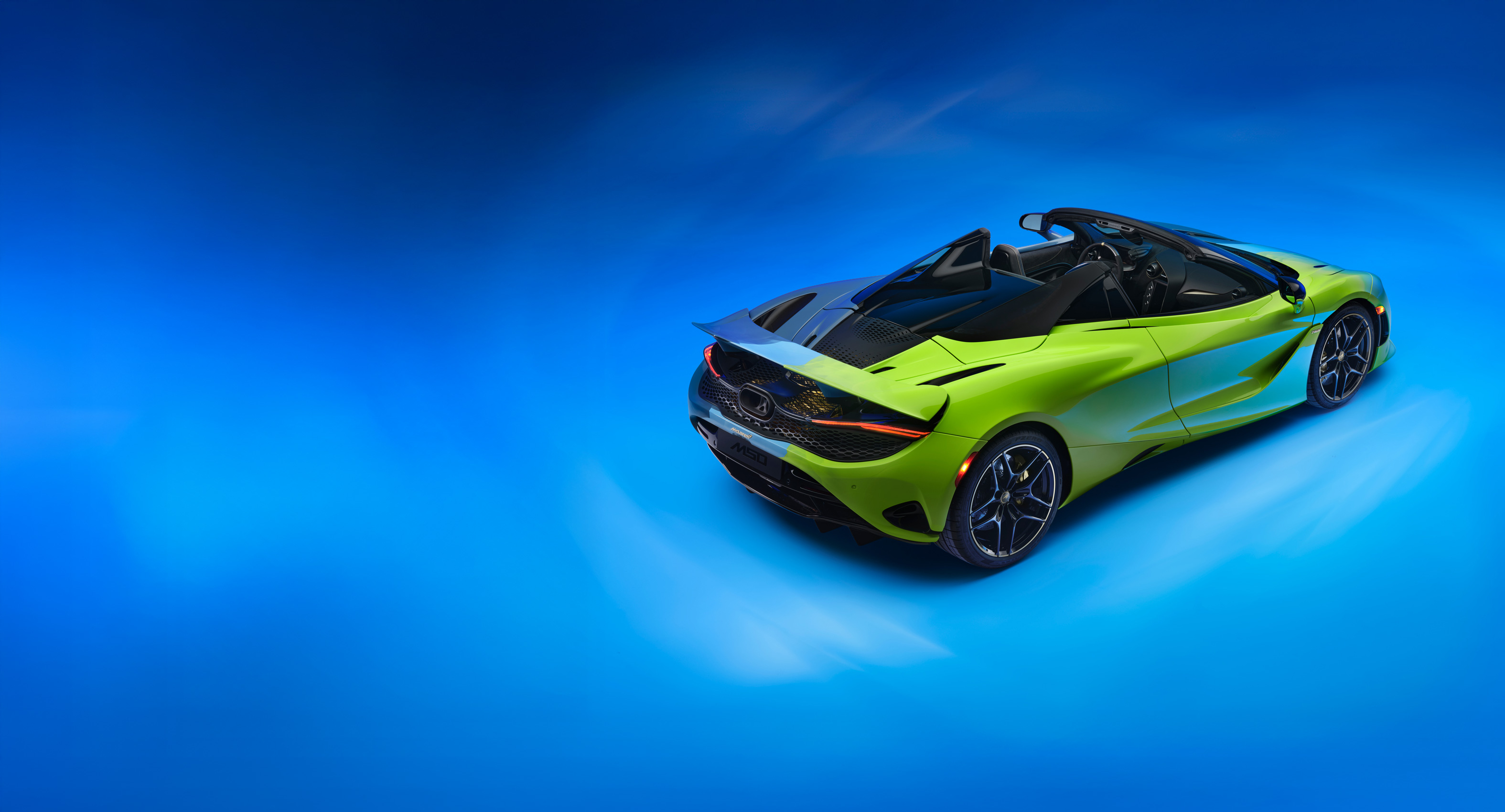 Rear of the Project Chromology 750s by MSO and Nat Bowen, on display against a glowing blue background. The car is various overlaying shades of green and blue and can be seen parked at an angle facing away from the camera.