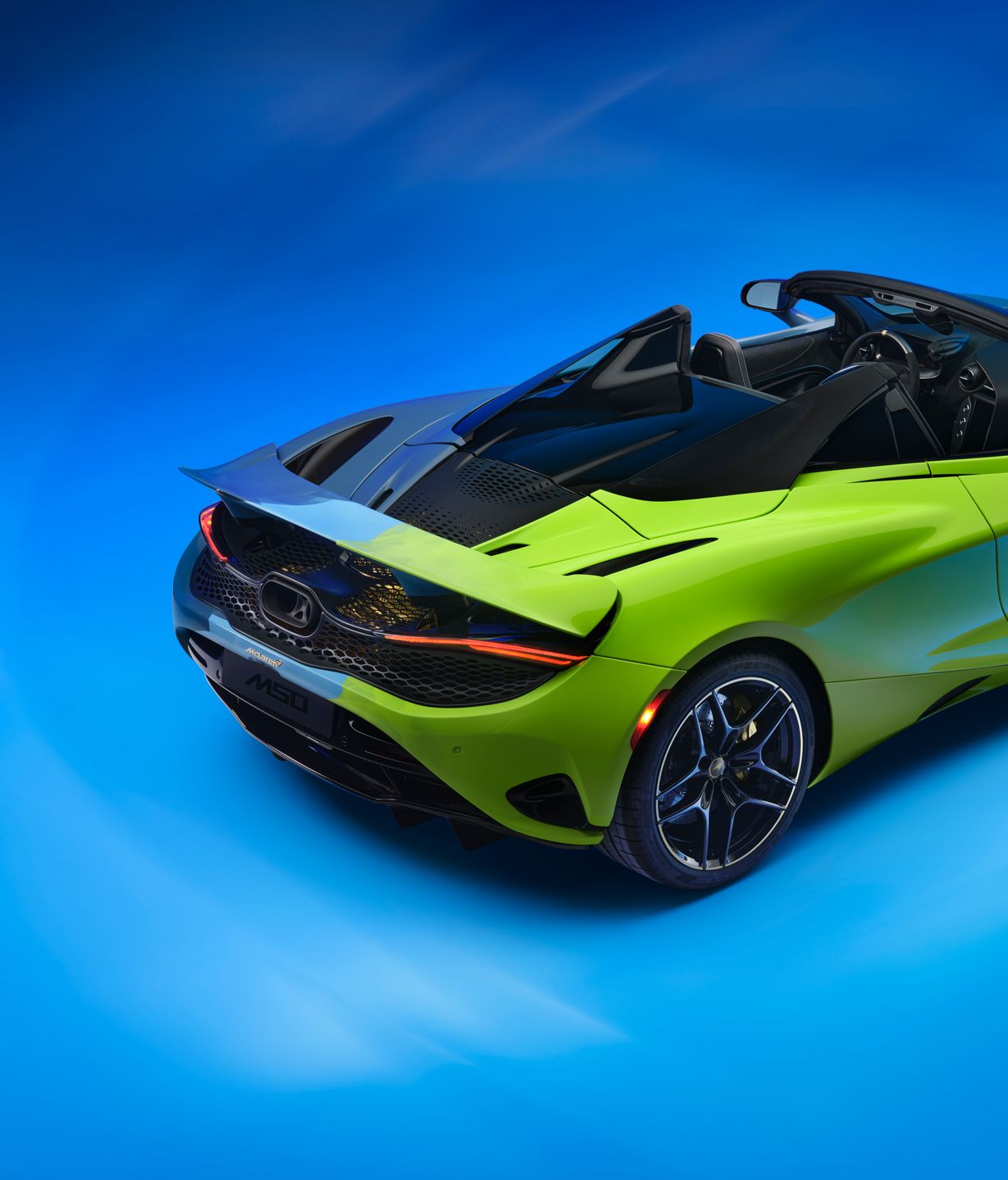 The rear side of the Project Chromology 750s by MSO and Nat Bowen, on display against a glowing blue background. The car is various overlaying shades of green and blue and can be seen parked at an angle with its headlights on.